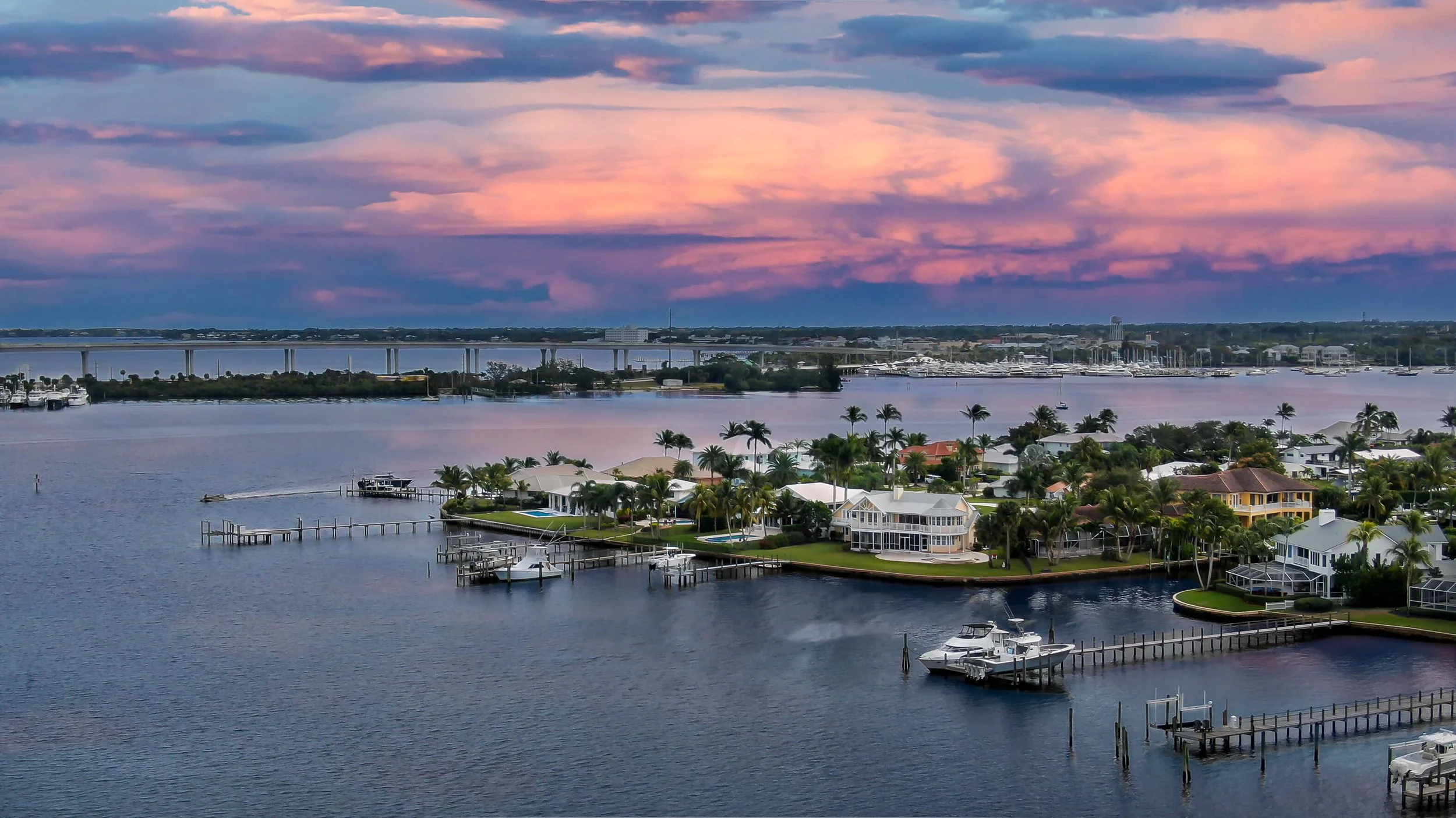A scenic view of a waterside neighborhood with houses, palm trees, and boats docked at piers during sunset, with a bridge and cityscape in the background.