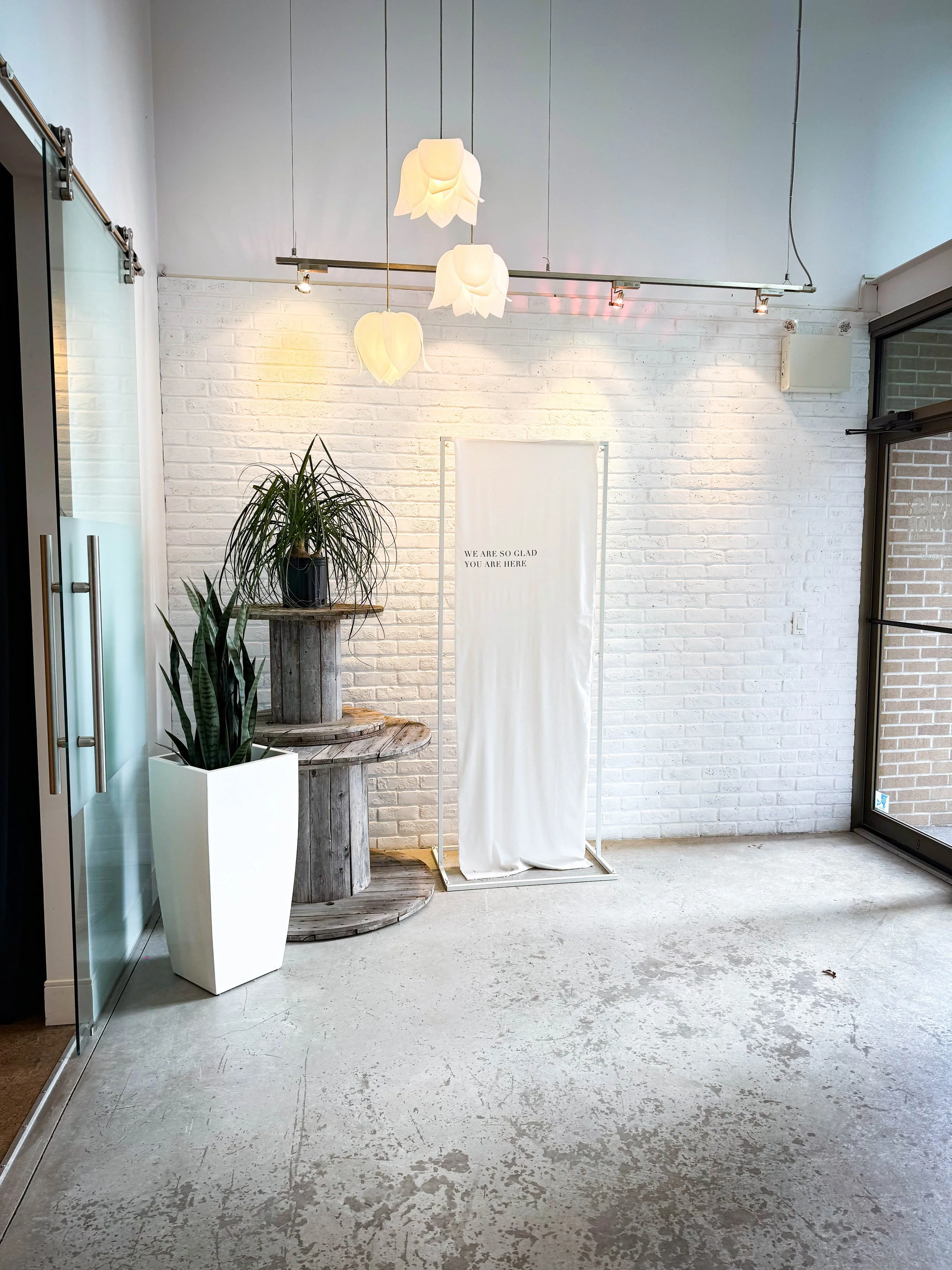Minimalist interior with white brick wall, three hanging ceiling lights resembling flowers, a potted plant on a wooden cable spool table, a tall white planter with a snake plant, and a white cloth standing banner with text, near glass door and window.