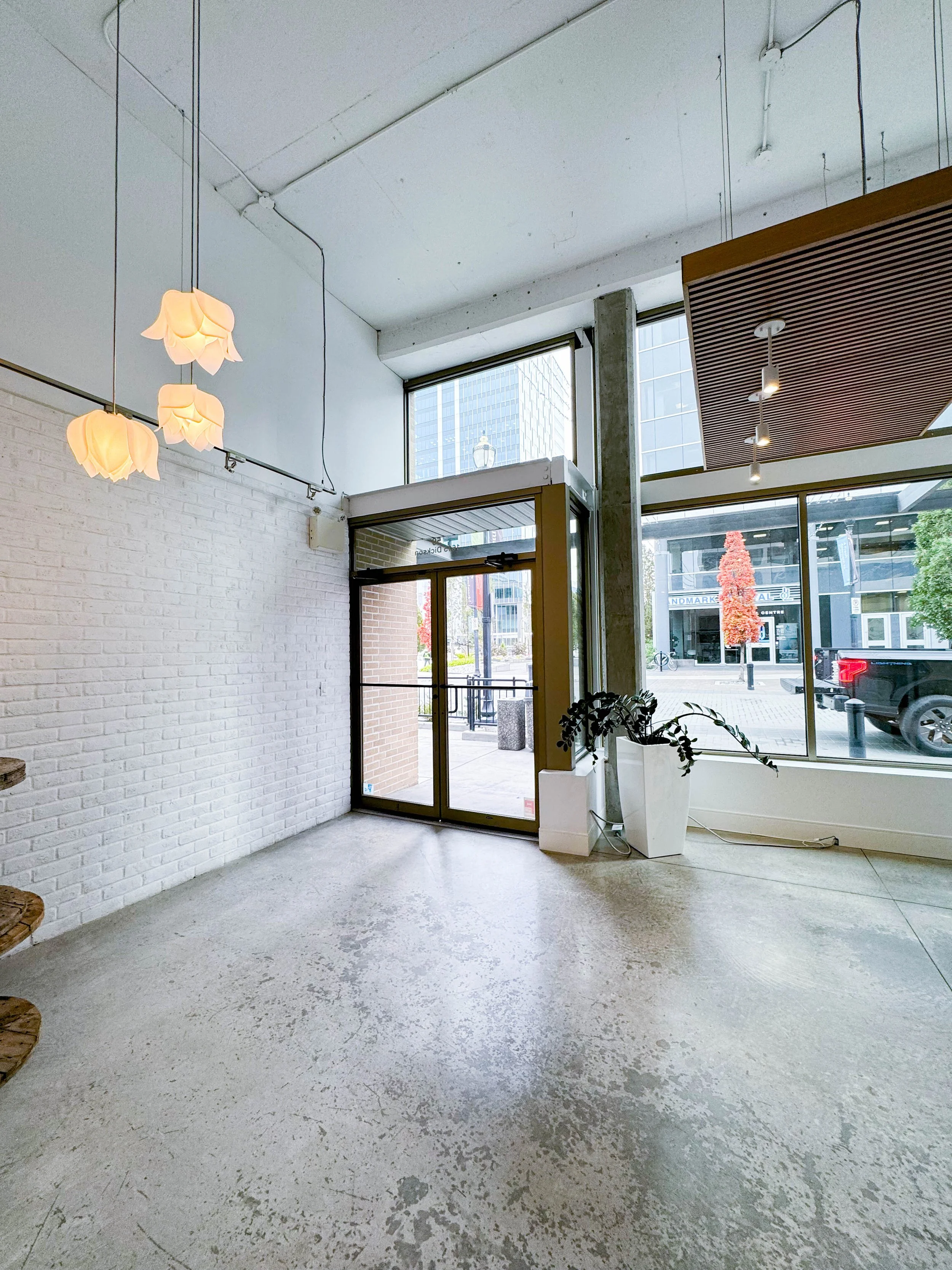 Empty store with white brick walls, large glass windows, hanging floral-shaped lights, and potted plants near the entrance.