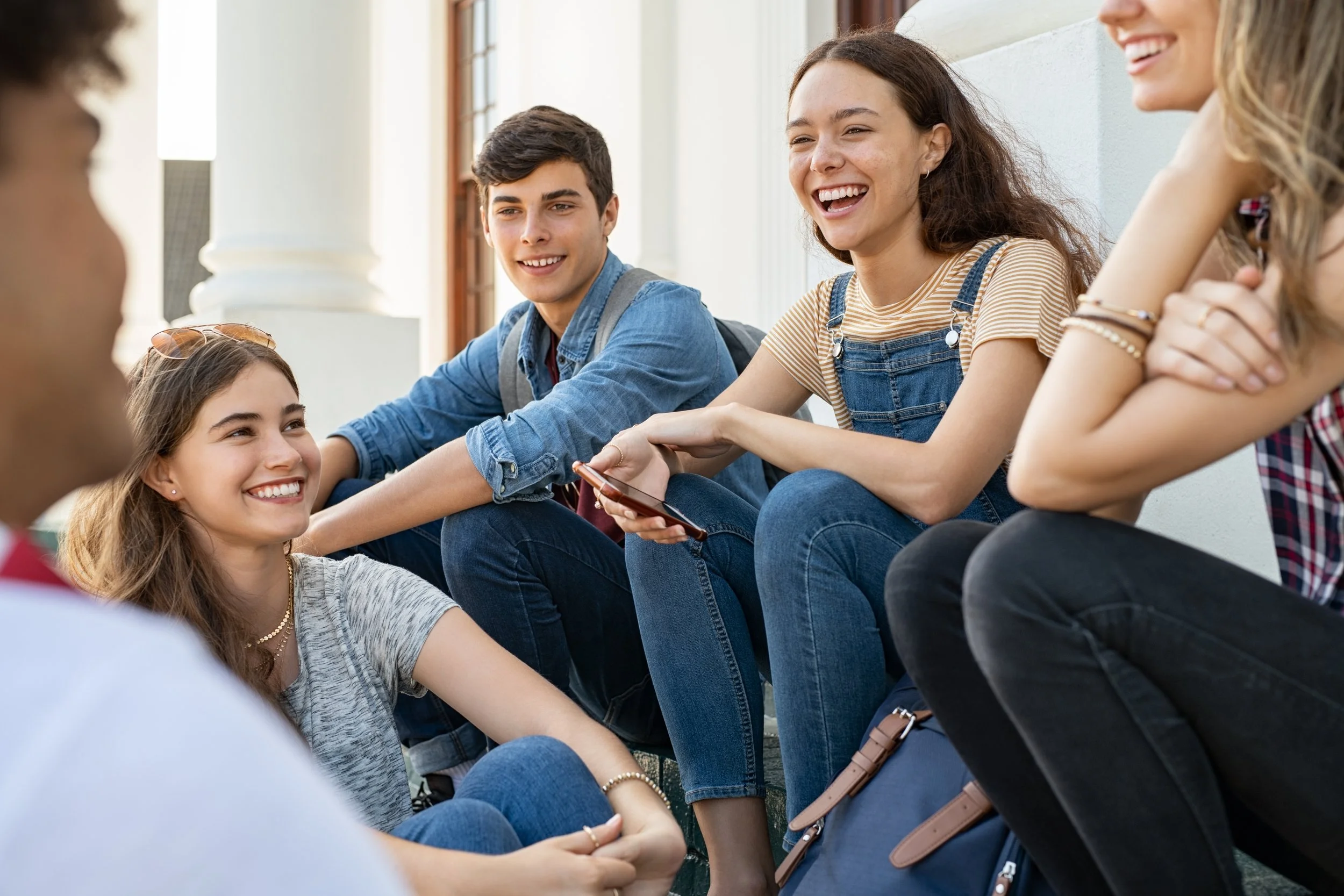 Group of five teenagers sitting on steps outside, smiling and talking, with one girl holding a phone.