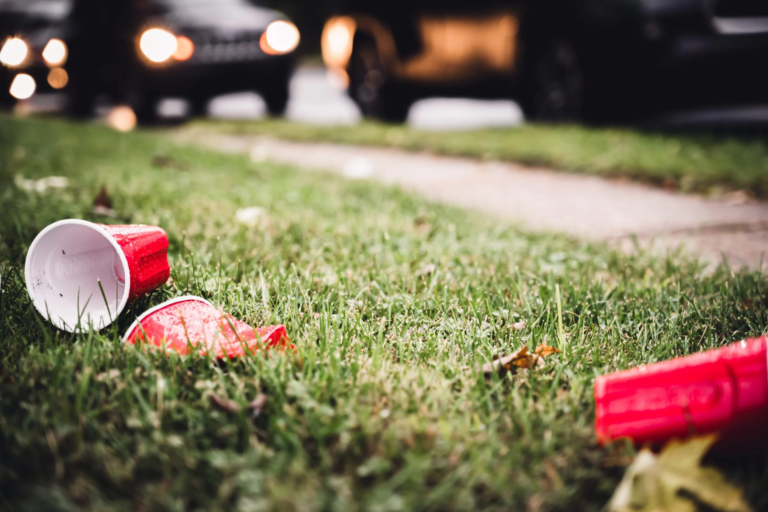 Littered red solo cups and cans scattered on grass near a sidewalk with cars in the background.