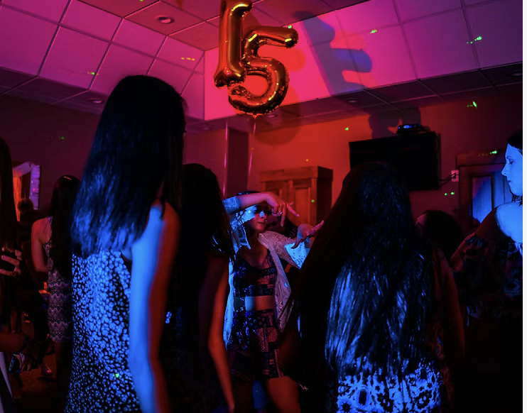 People dancing at a party under colorful lights with a large gold '15' balloon hanging from the ceiling.