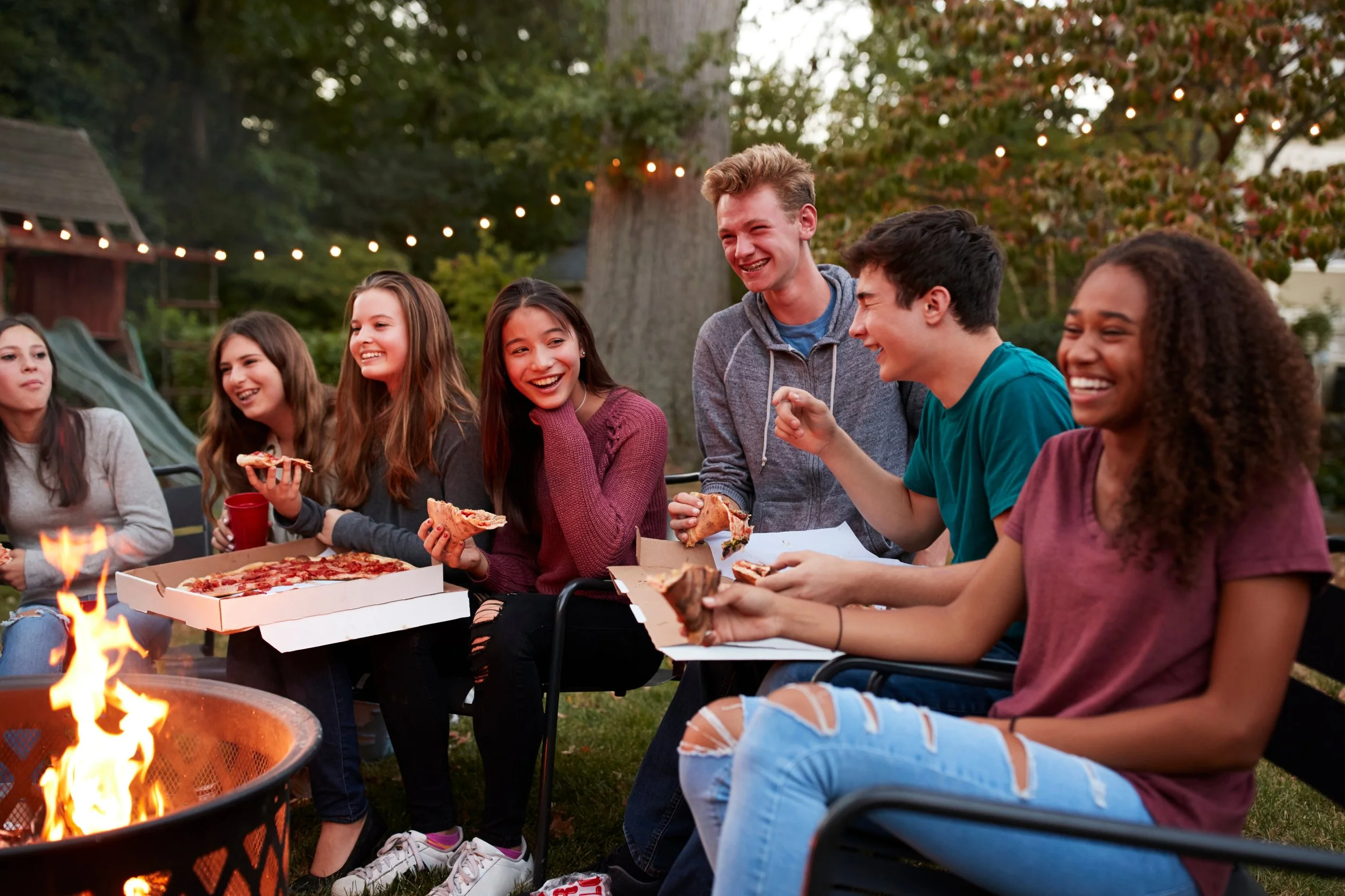 Group of teenagers enjoying pizza around a campfire outdoors during evening with string lights overhead.