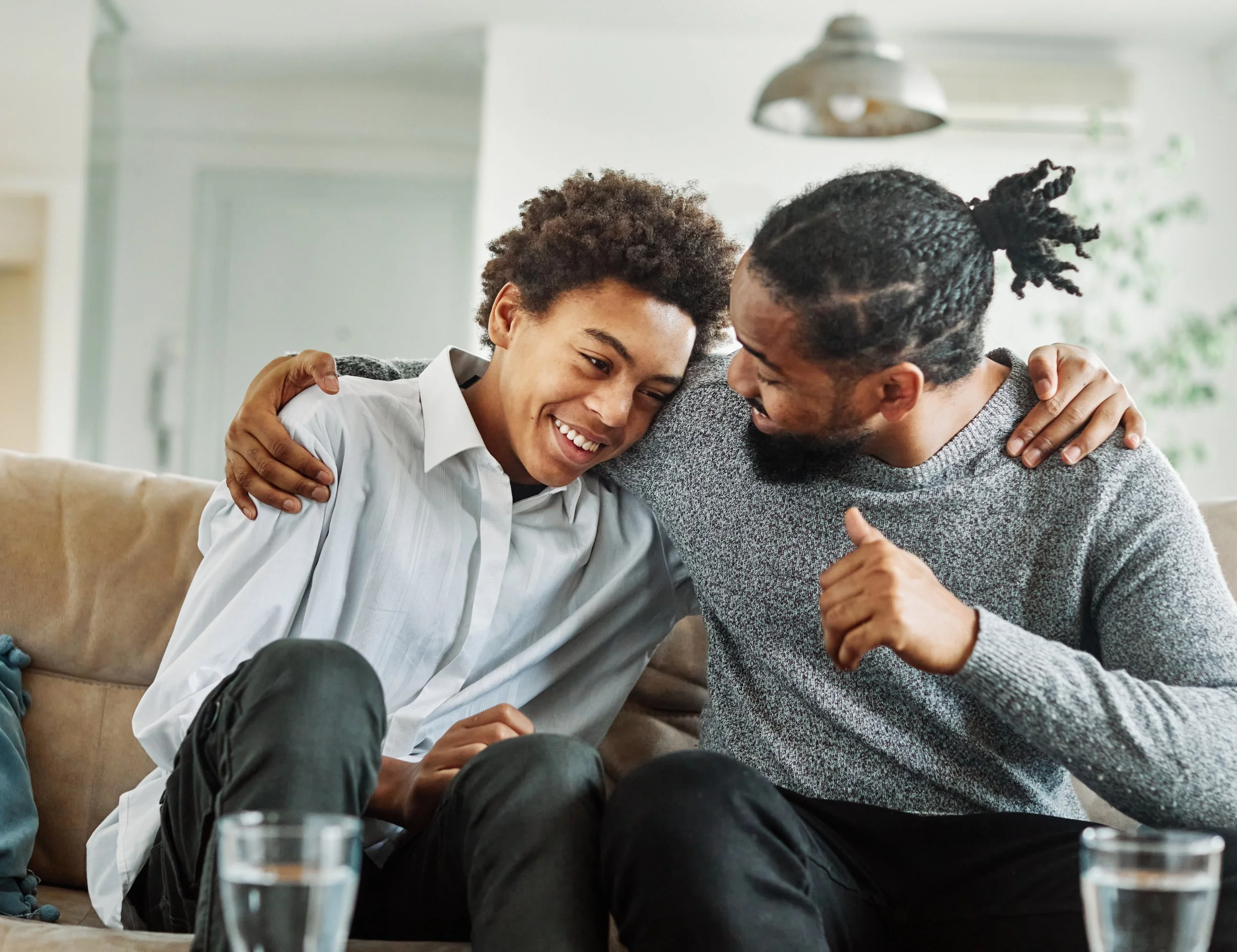 A young man and his father sit on a couch, smiling and laughing together, with their arms around each other in a cozy living room.