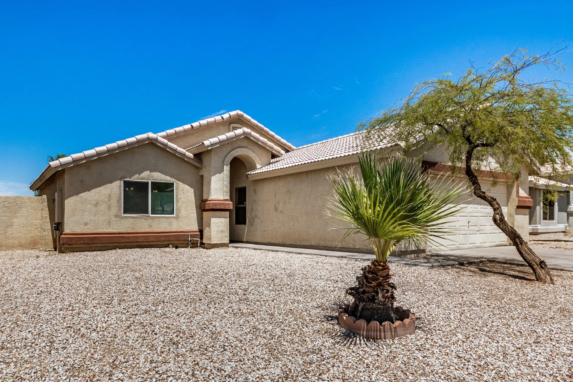 Front view of a beige single-story house with a gravel yard, a small palm tree in a circular planter, another tree to the right, and a blue sky overhead.