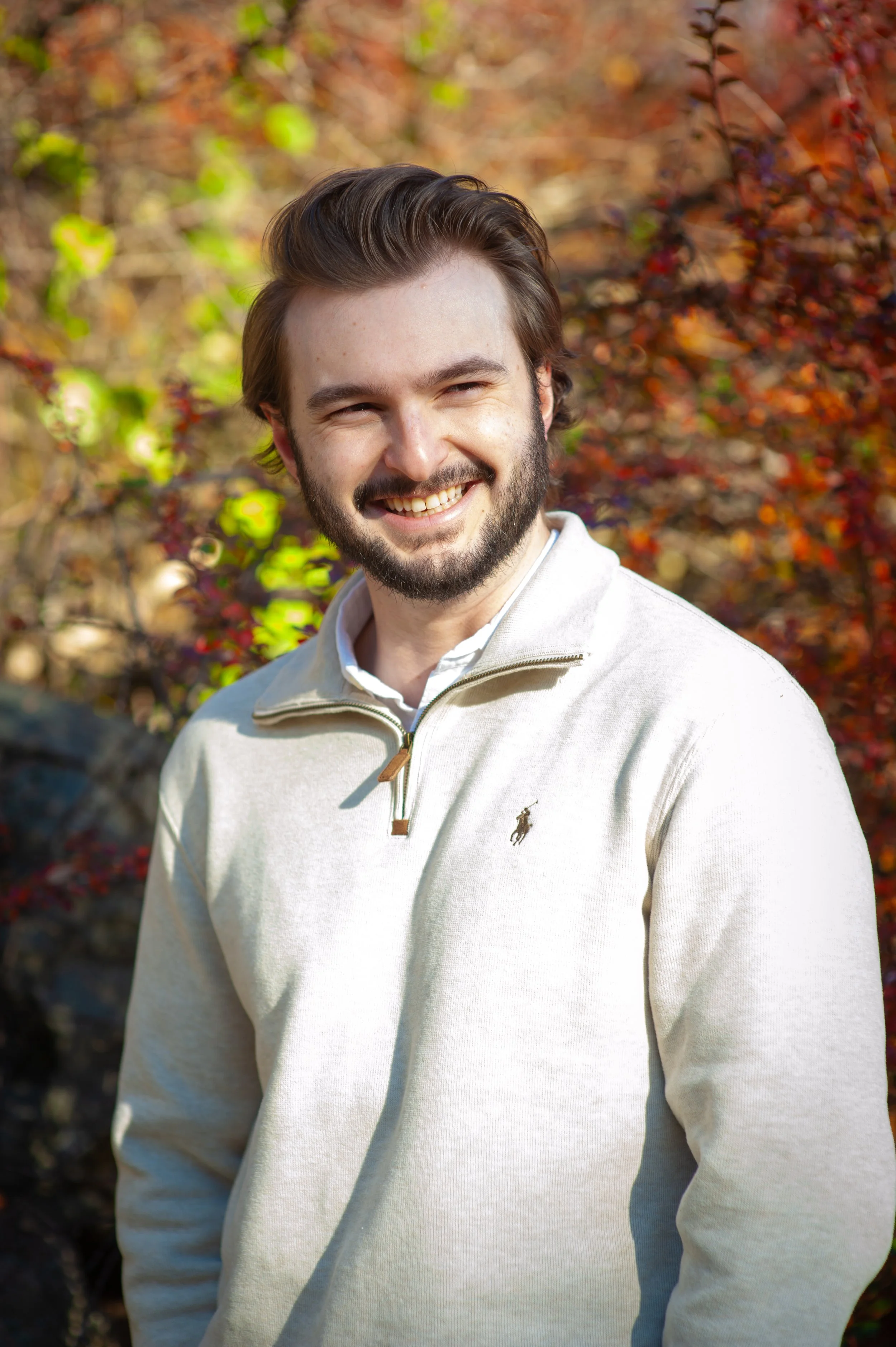 A smiling man with dark hair and a beard, wearing a light-colored quarter-zip sweater, standing outdoors with autumn foliage in the background.