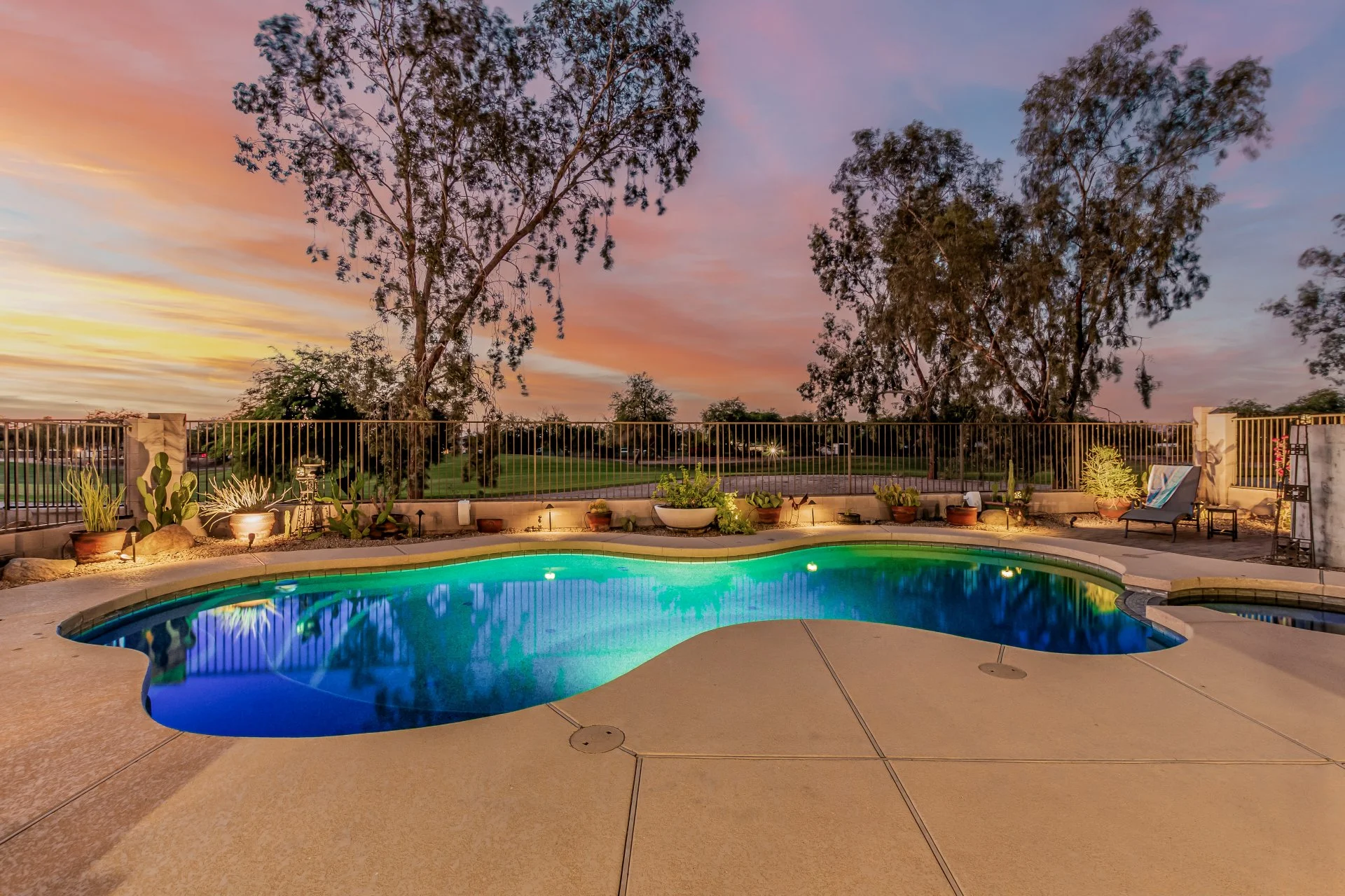 A backyard swimming pool illuminated at dusk with sunset sky, surrounded by potted plants, a lounge chair, and outdoor decorative items.