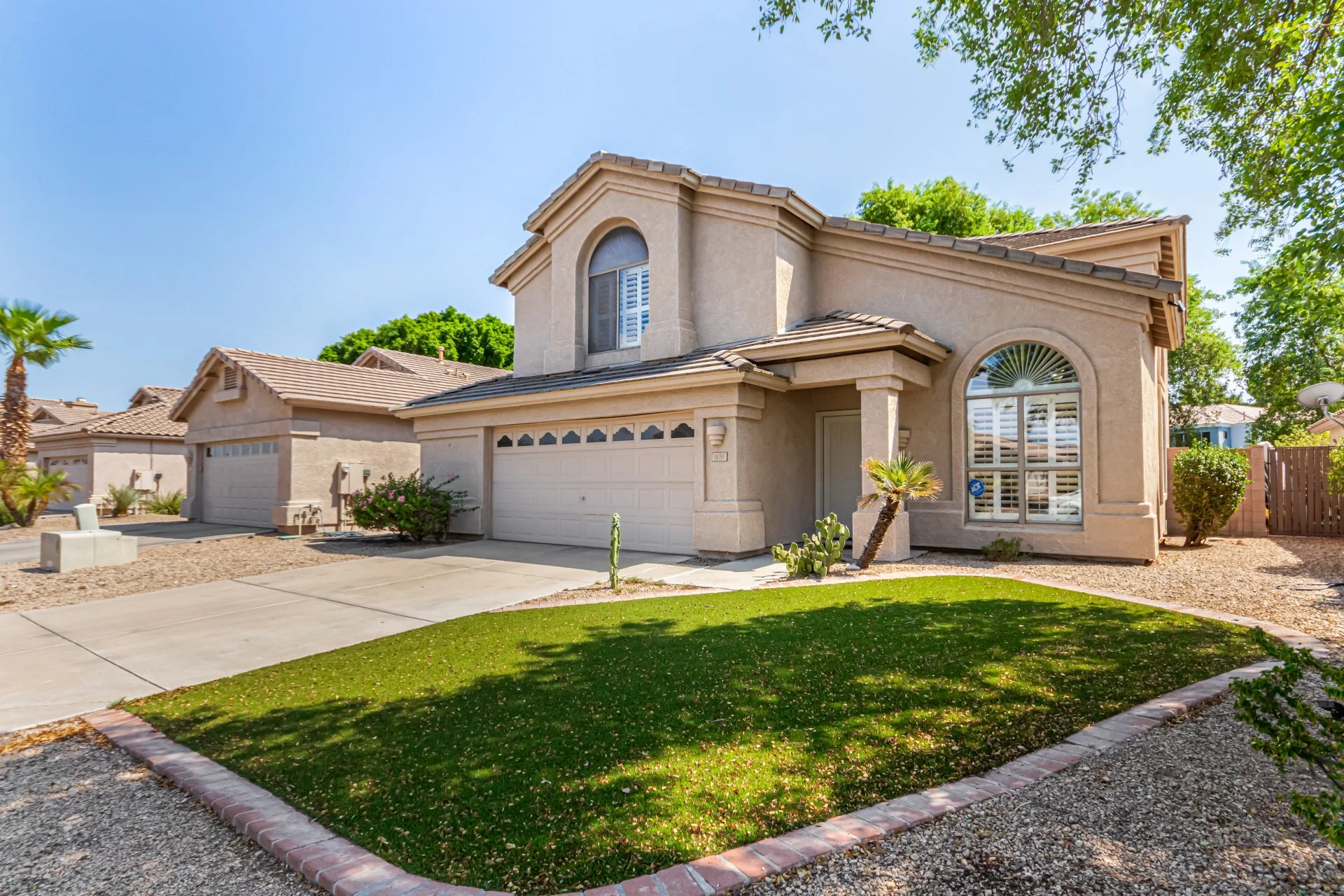 A two-story beige house with a well-maintained front yard featuring a small grass patch, desert landscaping, and a palm tree. The house has a white double garage door, arched window, and shutters.