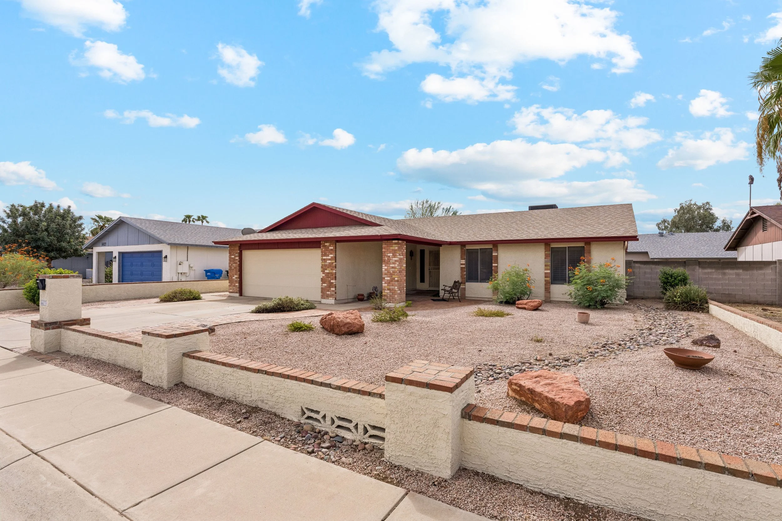 Front view of a suburban house with a brick and stucco exterior, two-car garage, and a small front porch, surrounded by a low brick wall, with desert landscaping and a sidewalk in the foreground under a blue sky with white clouds.