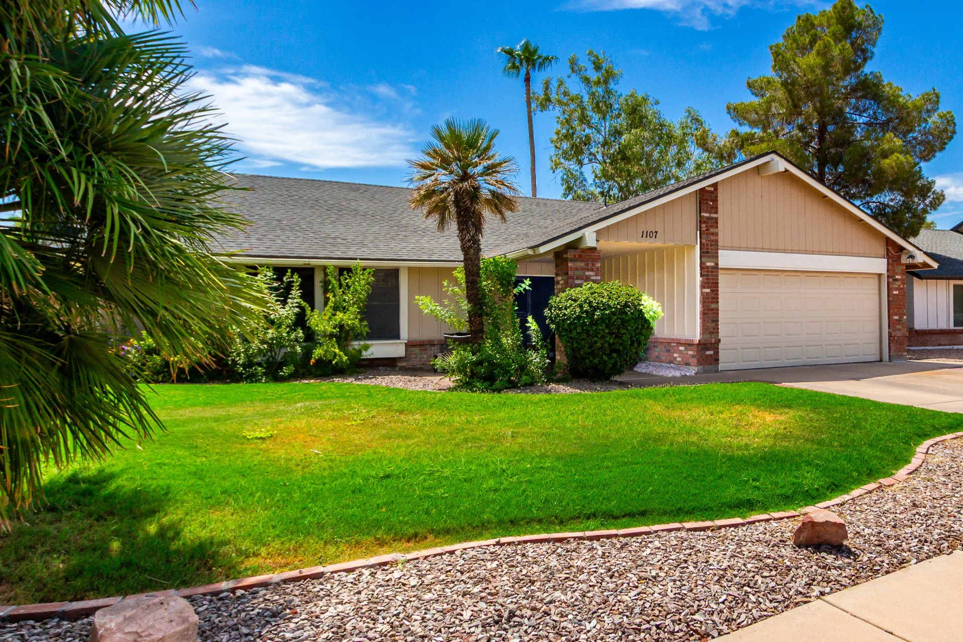 Front view of a single-story suburban house with a green lawn, bushes, and trees, including a palm tree, under a blue sky.