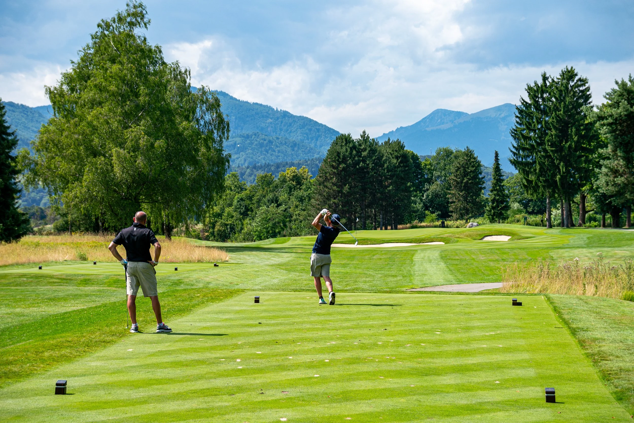 Due uomini che giocano a golf su un campo con montagne sullo sfondo, uno che sta per colpire la palla e l'altro che osserva.