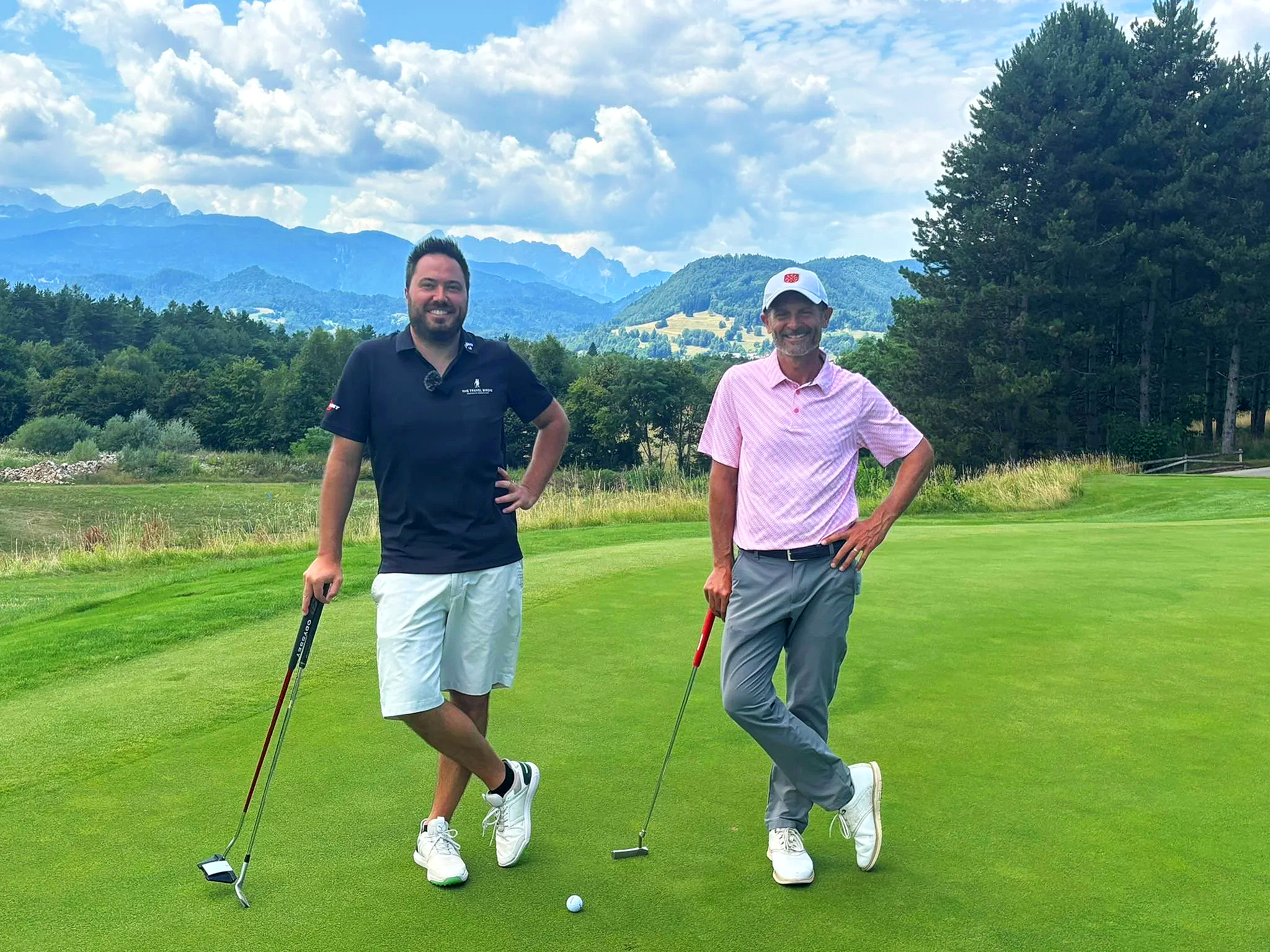 Due uomini che giocano a golf su un campo verde con montagne sullo sfondo, uno con maglietta nera e pantaloncini bianchi, l'altro con camicia rosa e pantaloni grigi.