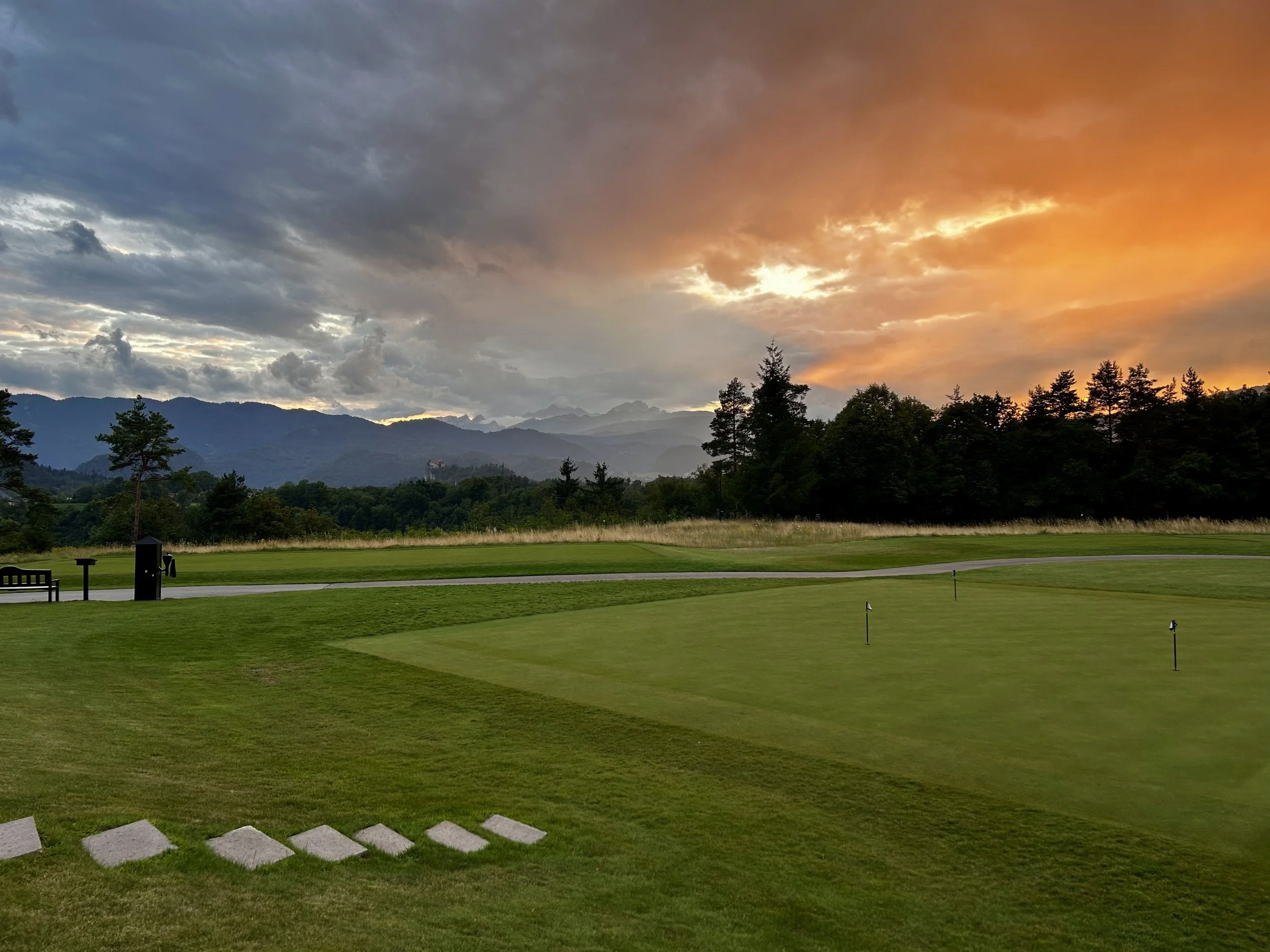 Campo da golf con verde e bandiere, al tramonto con cielo arancione e nuvole, montagne sullo sfondo e alberi lungo il bordo del campo.