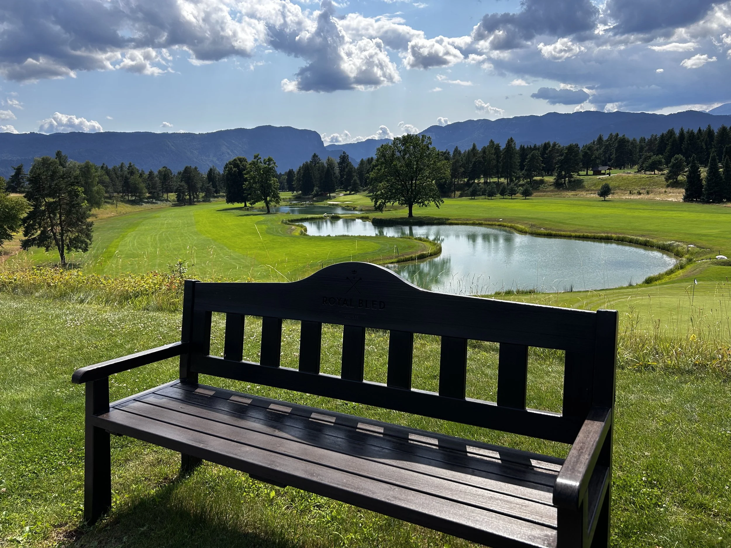 Panorama di un campo da golf con alberi, un laghetto, montagne sullo sfondo e un cielo con nuvole, vista da una panchina di legno sulla erba.