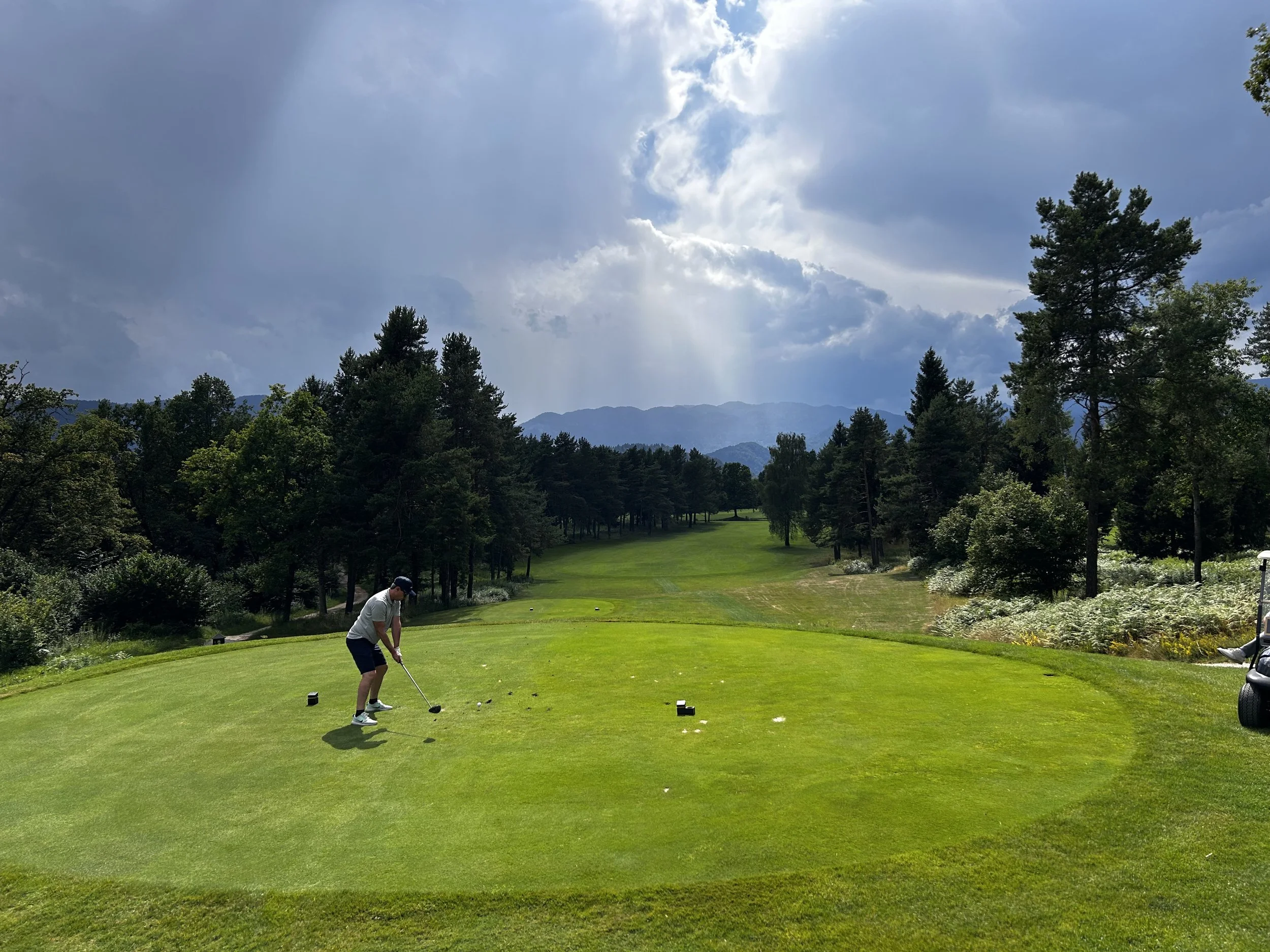 Giocatore di golf che si prepara a colpire la pallina su un fairway di un campo da golf immerso nella natura con alberi e montagne sullo sfondo, sotto un cielo parzialmente nuvoloso.