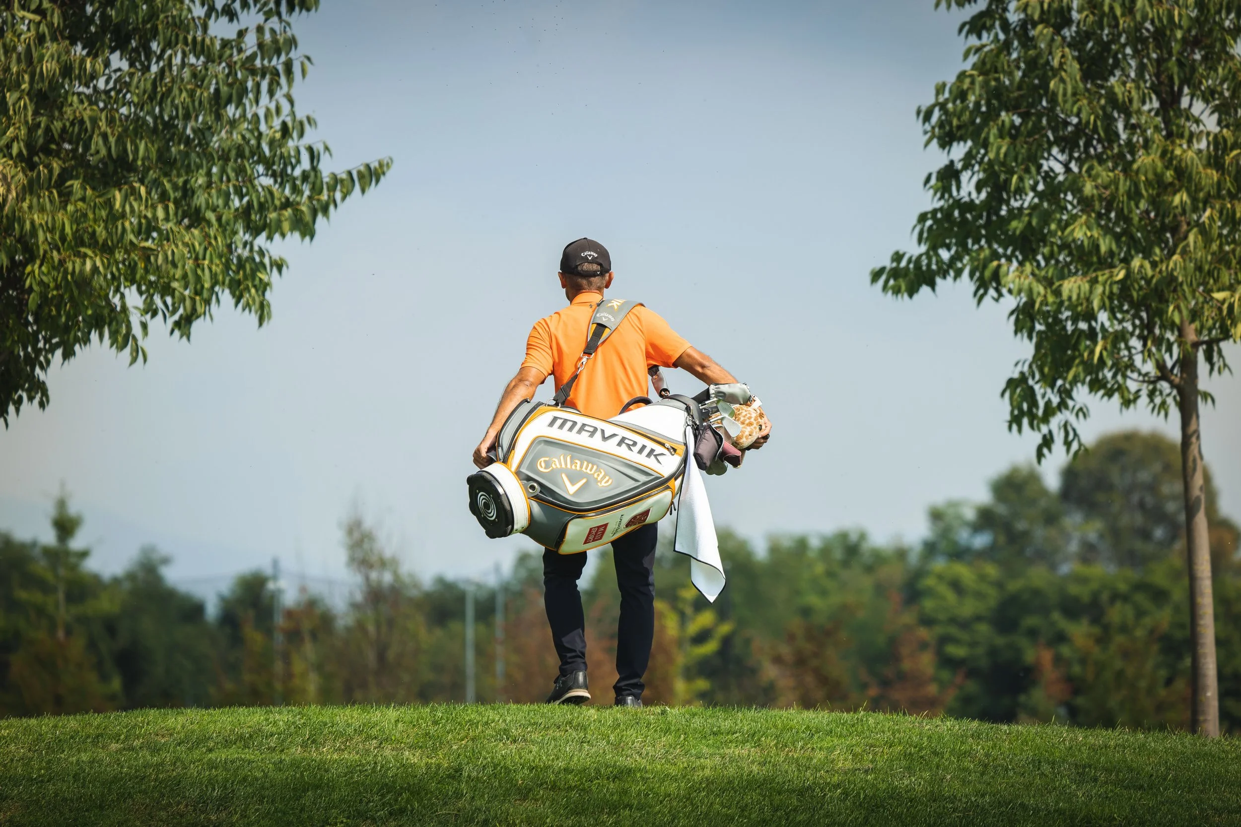 Un uomo con cappellino, in maglietta arancione, che porta una borsa da golf con il marchio Callaway, sta camminando su un campo di golf tra gli alberi, con un cielo sereno.