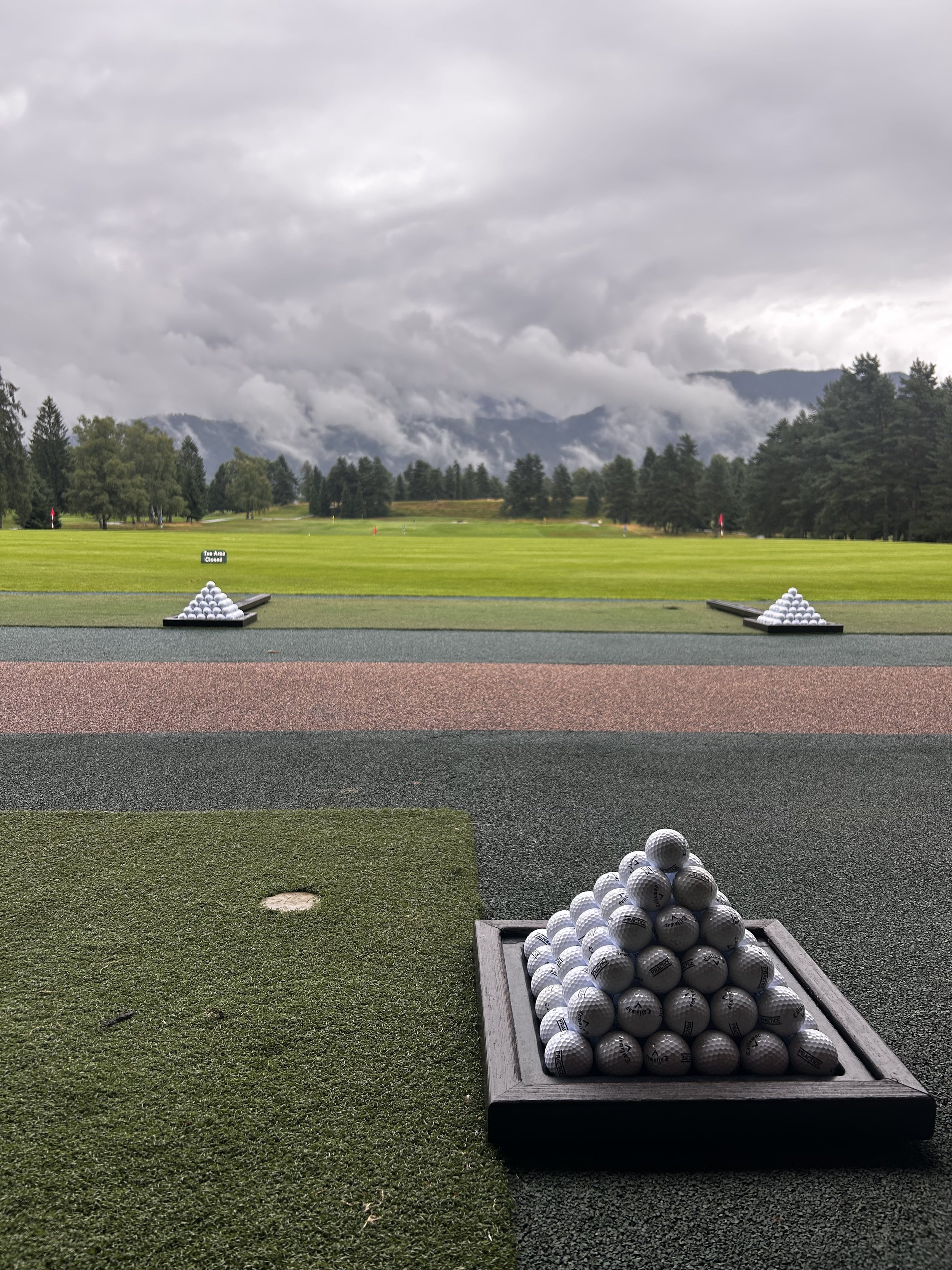 Ceste da golf con palle di golf impilate, in un campo da golf sotto un cielo nuvoloso, con alberi in lontananza e montagne sullo sfondo.