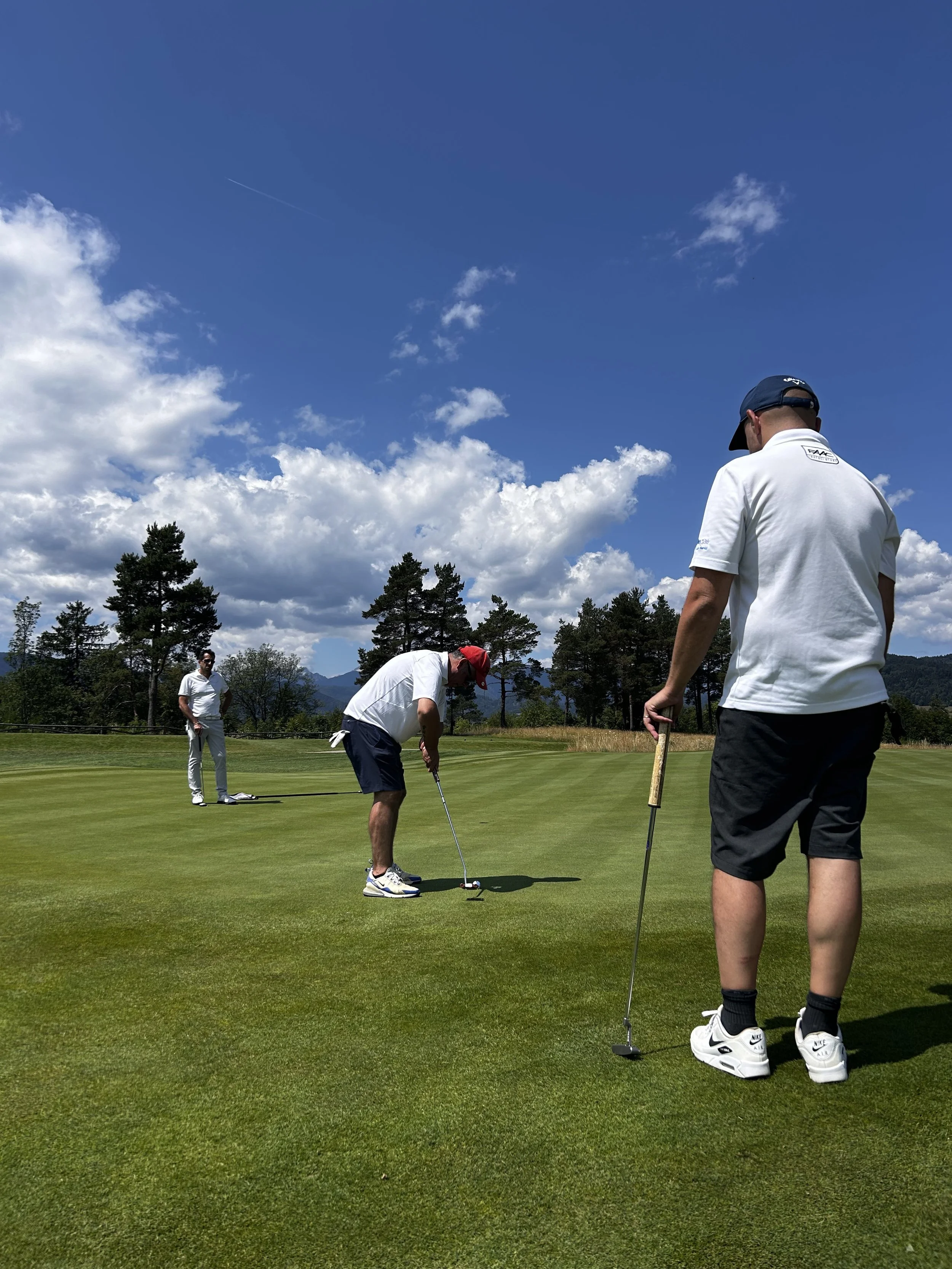 Tre persone che giocano a golf su un campo con alberi e montagne sullo sfondo, sotto un cielo blu con nuvole.