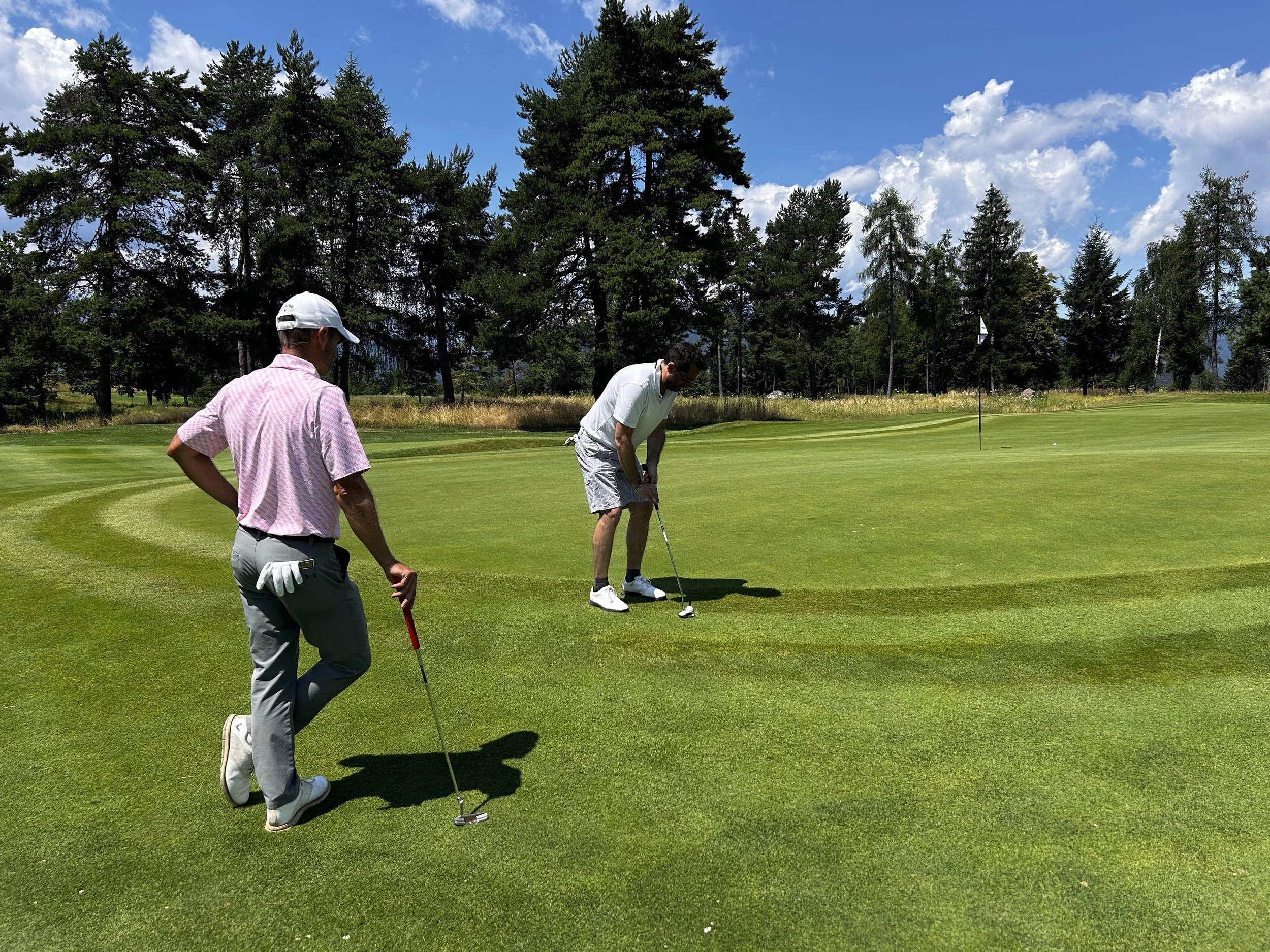 Due uomini che giocano a golf su un campo ben curato, uno che si sta preparando a mettere la palla vicino alla buca, mentre l'altro osserva. Sullo sfondo ci sono alberi e un cielo nuvoloso.