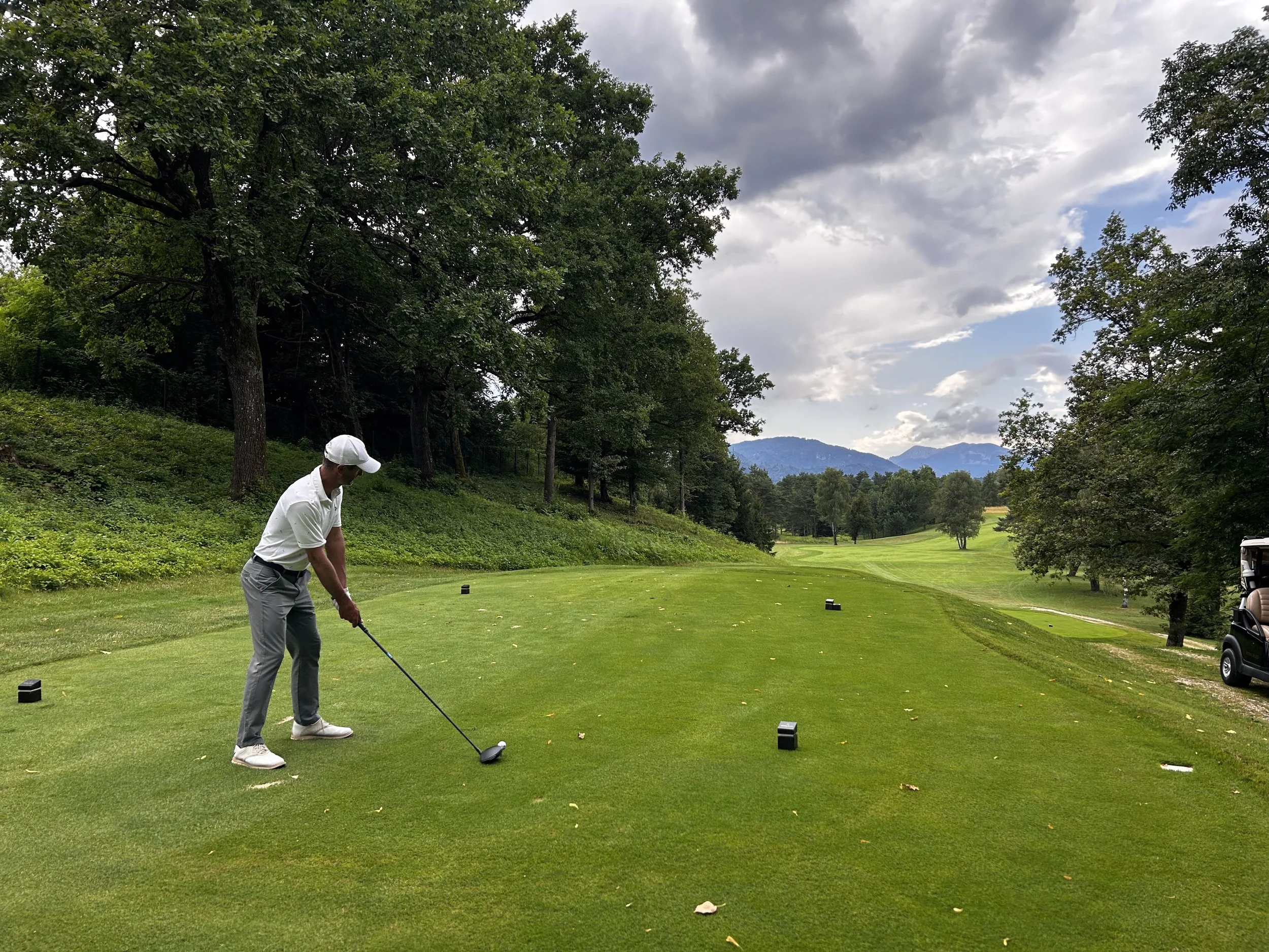 Un uomo che gioca a golf su un campo verde circondato da alberi e montagne in lontananza, con nuvole nel cielo.