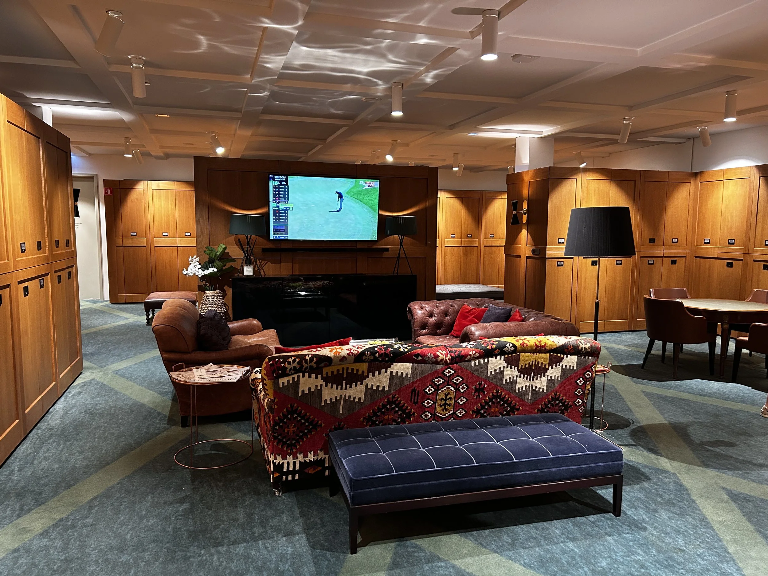 Uno spazio lounge con divani e poltrone, una grande televisione che mostra un golfista in campo, pareti rivestite di pannelli di legno, lampade nere, un tavolo rotondo con sedie marroni e un tappeto con disegni geometrici.