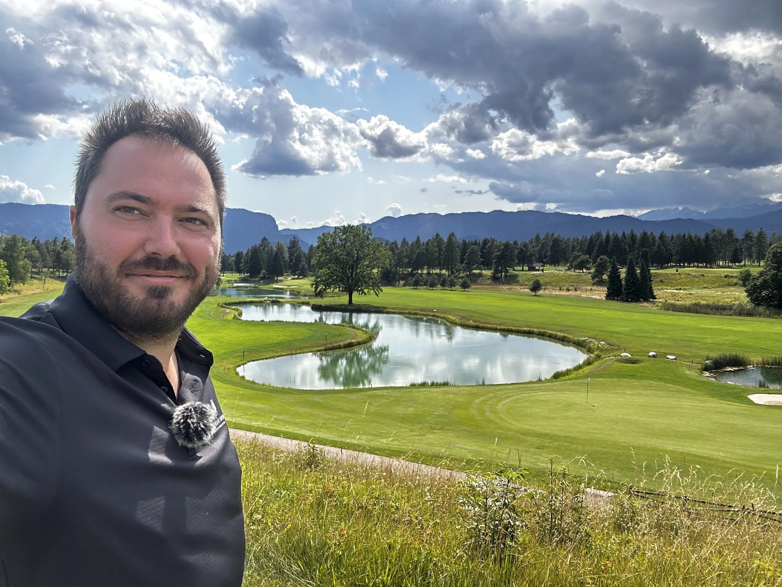 Uomo che si trova su un campo da golf con laghetto, alberi e montagne sullo sfondo, sotto un cielo in parte nuvoloso.