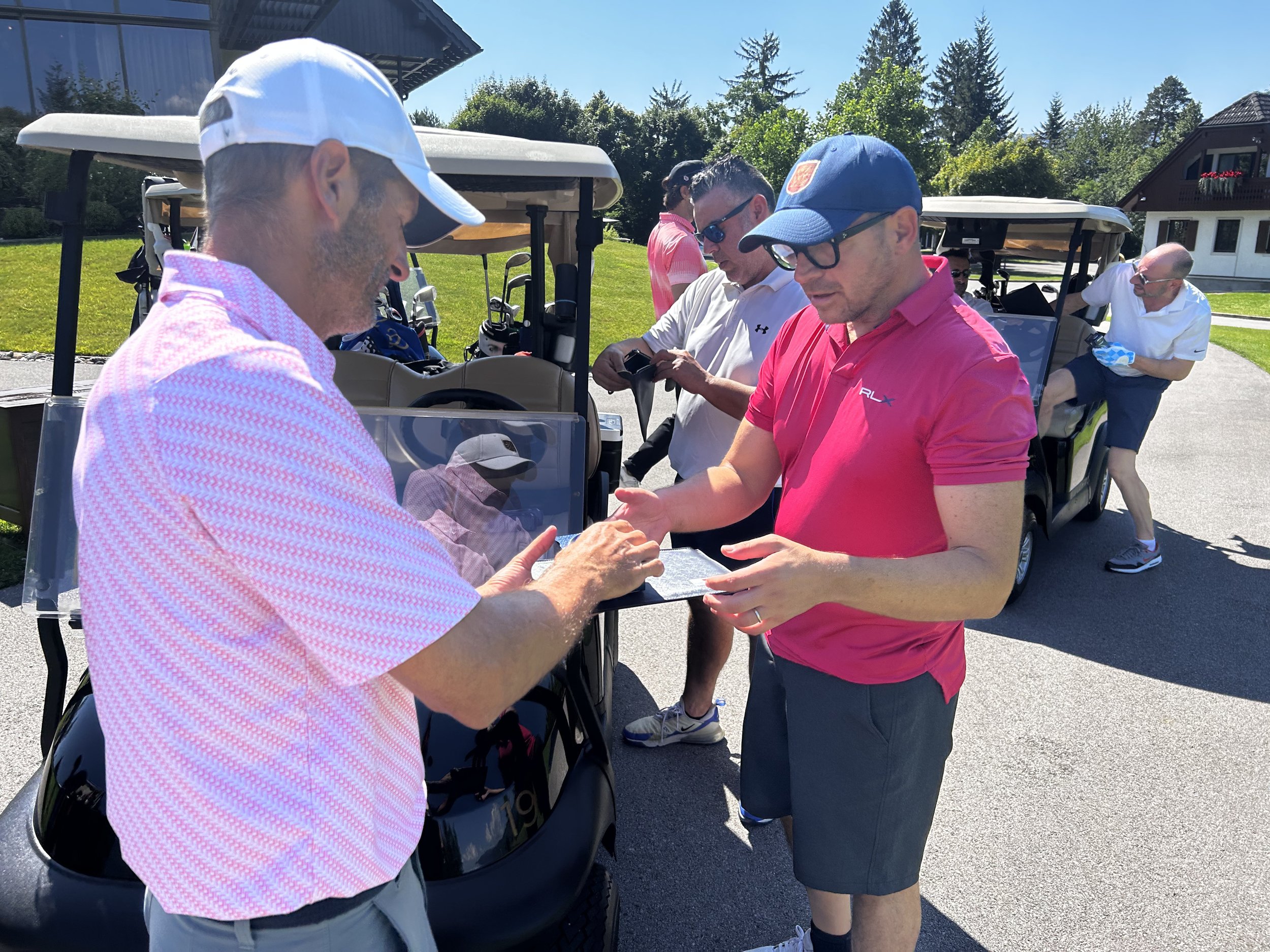 Gruppo di persone che si scambiano qualcosa su un campo da golf, con golf cart sullo sfondo, in un'area verde soleggiata