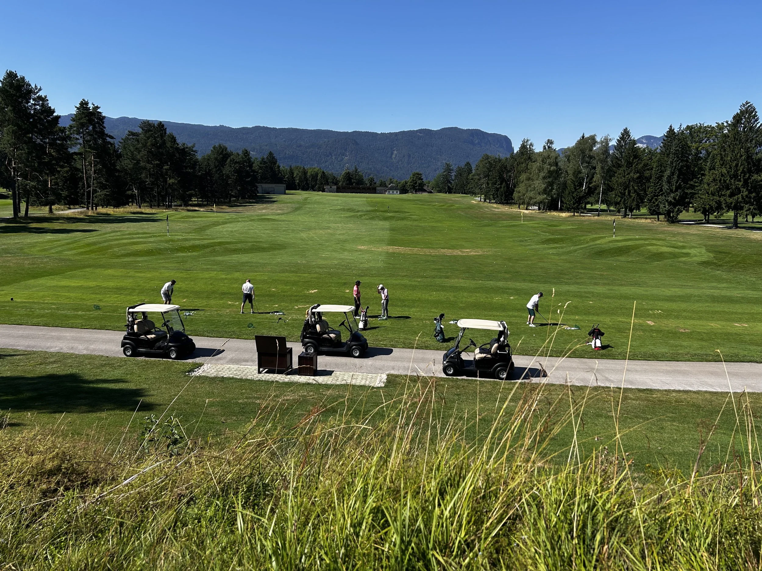 Campo da golf con persone che giocano e vetturette da golf in un paesaggio verde con montagna sullo sfondo.
