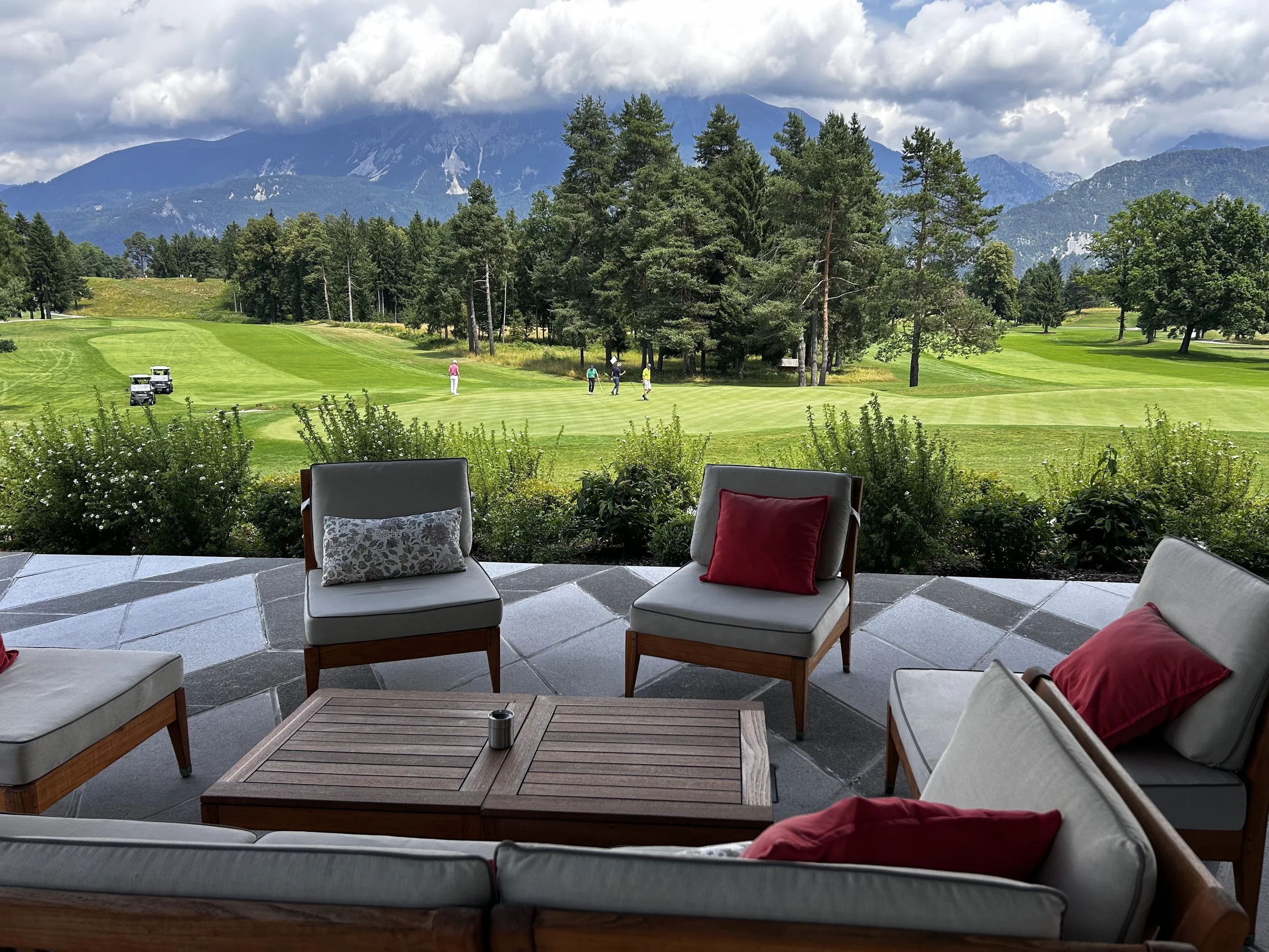 Vista su un campo da golf con persone che giocano, circondato da alberi e montagne sullo sfondo, con una zona di relax con sedie e tavolino in primo piano.