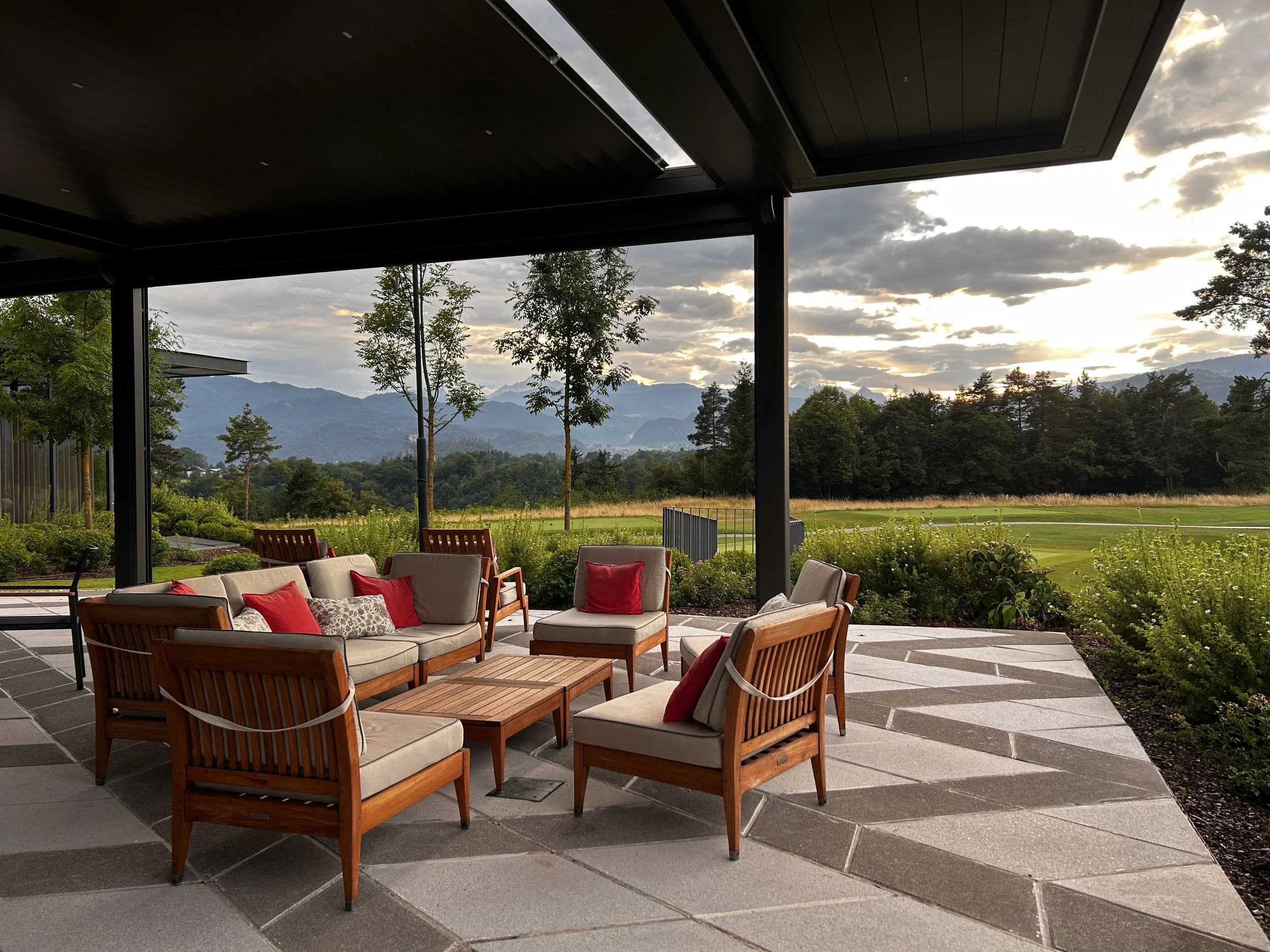 Zona esterna con mobili da giardino in legno con cuscini beige e cuscini rossi, sotto una tettoia, con vista su un paesaggio naturale con alberi, prato e montagne in lontananza, al tramonto.