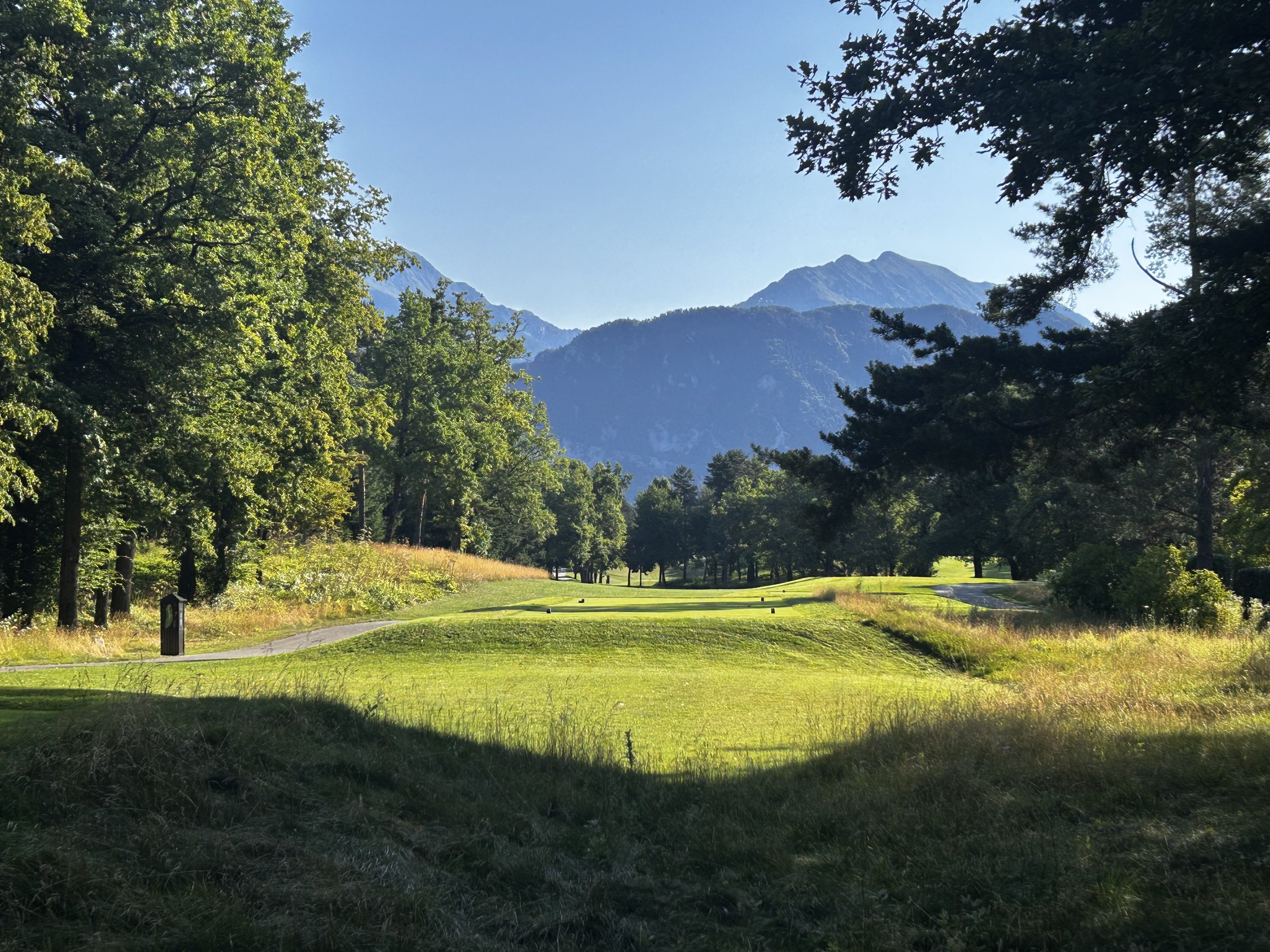 Campo di golf circondato da alberi e montagne sullo sfondo, con il cielo chiaro.