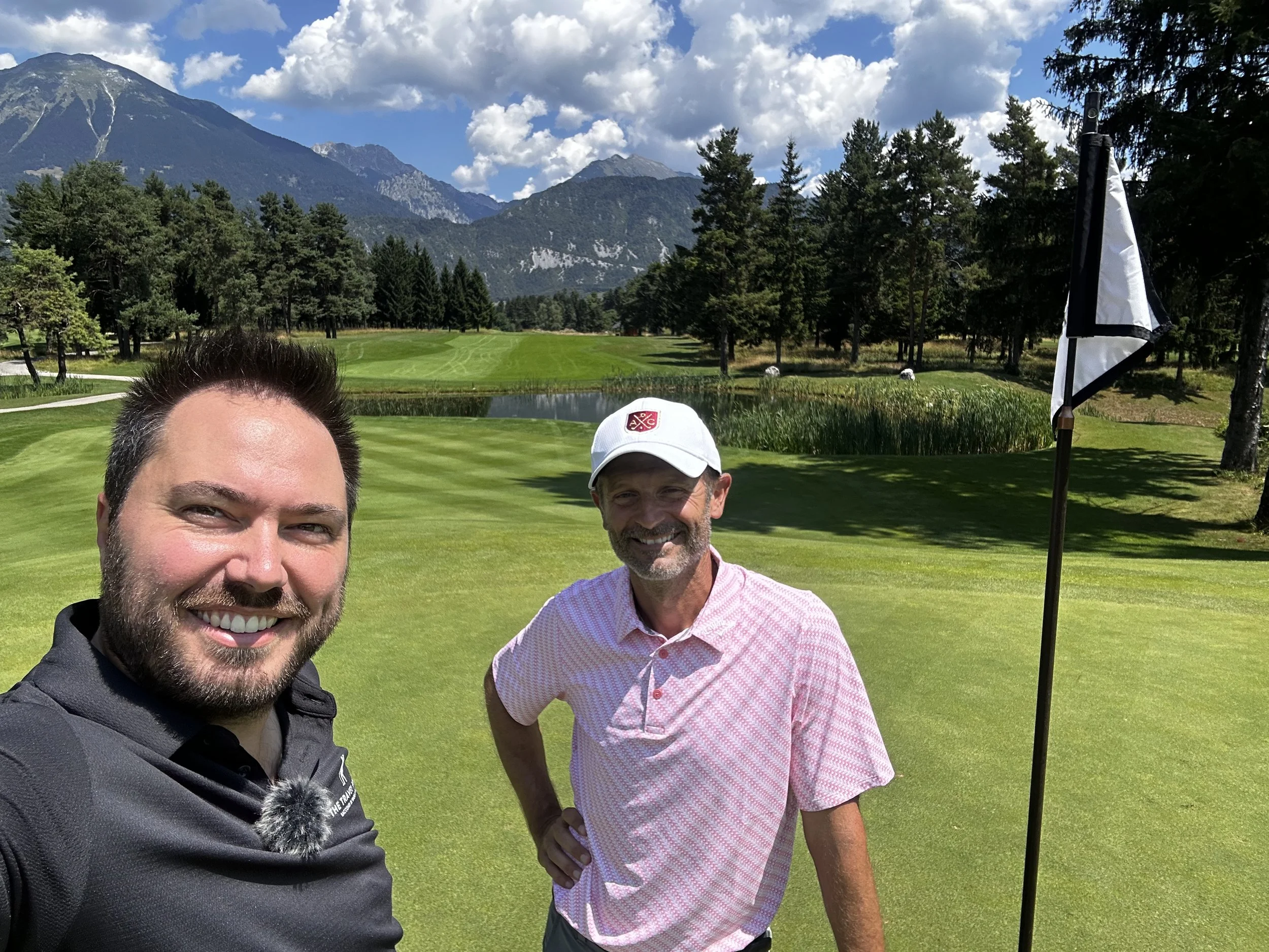 Due uomini sorridenti si trovano su un campo da golf con montagne sullo sfondo, un albero e una bandiera bianca e nera