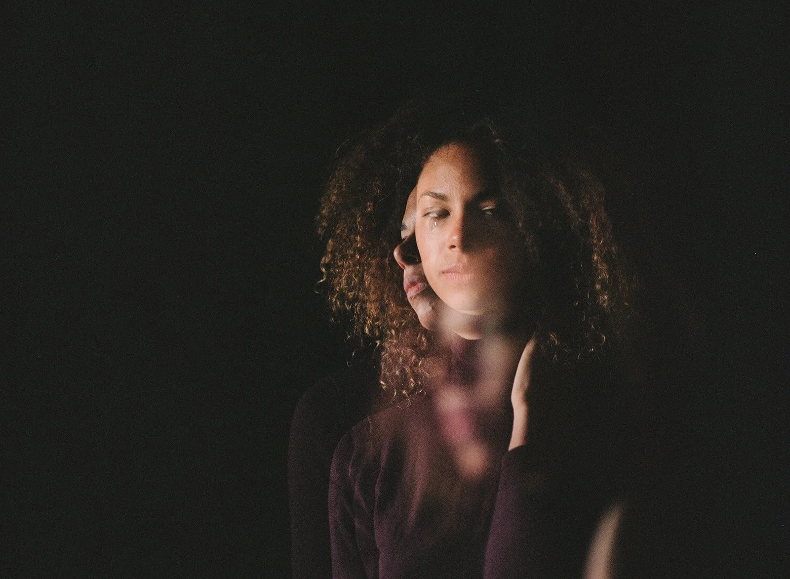 A woman with curly hair and a serious expression, with a faint reflection of her face overlapping on the left side, set against a dark background.