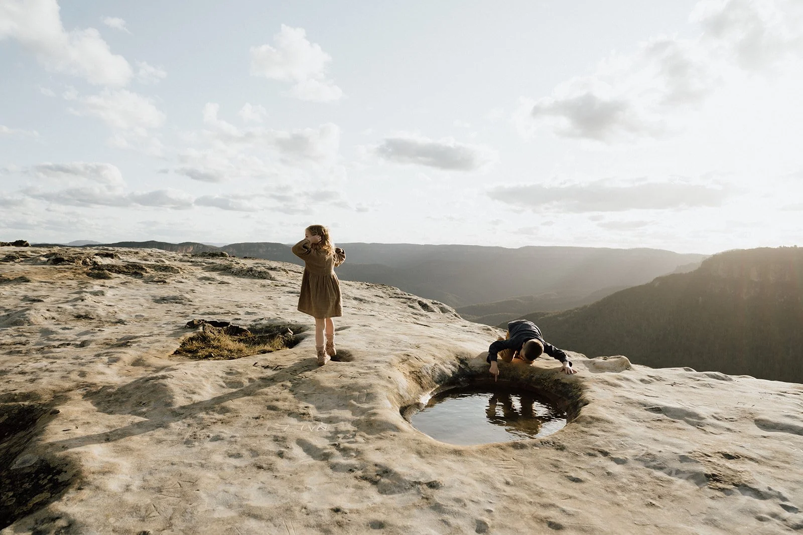 A girl in a beige coat standing next to a small water pool, and a boy kneeling at the edge drinking from the pool, on a rocky landscape with distant mountains and a cloudy sky at sunset.