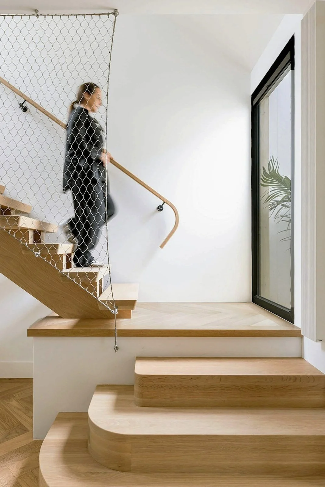 A woman walking down a modern wooden staircase with a safety net on one side, near a sliding glass door with a plant visible outside - interior photography, Sydney.