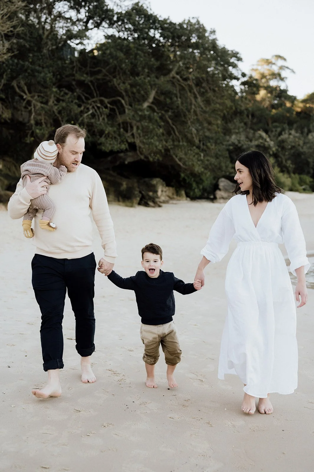 A family of four walking on a sandy beach, holding hands and smiling, with trees in the background.