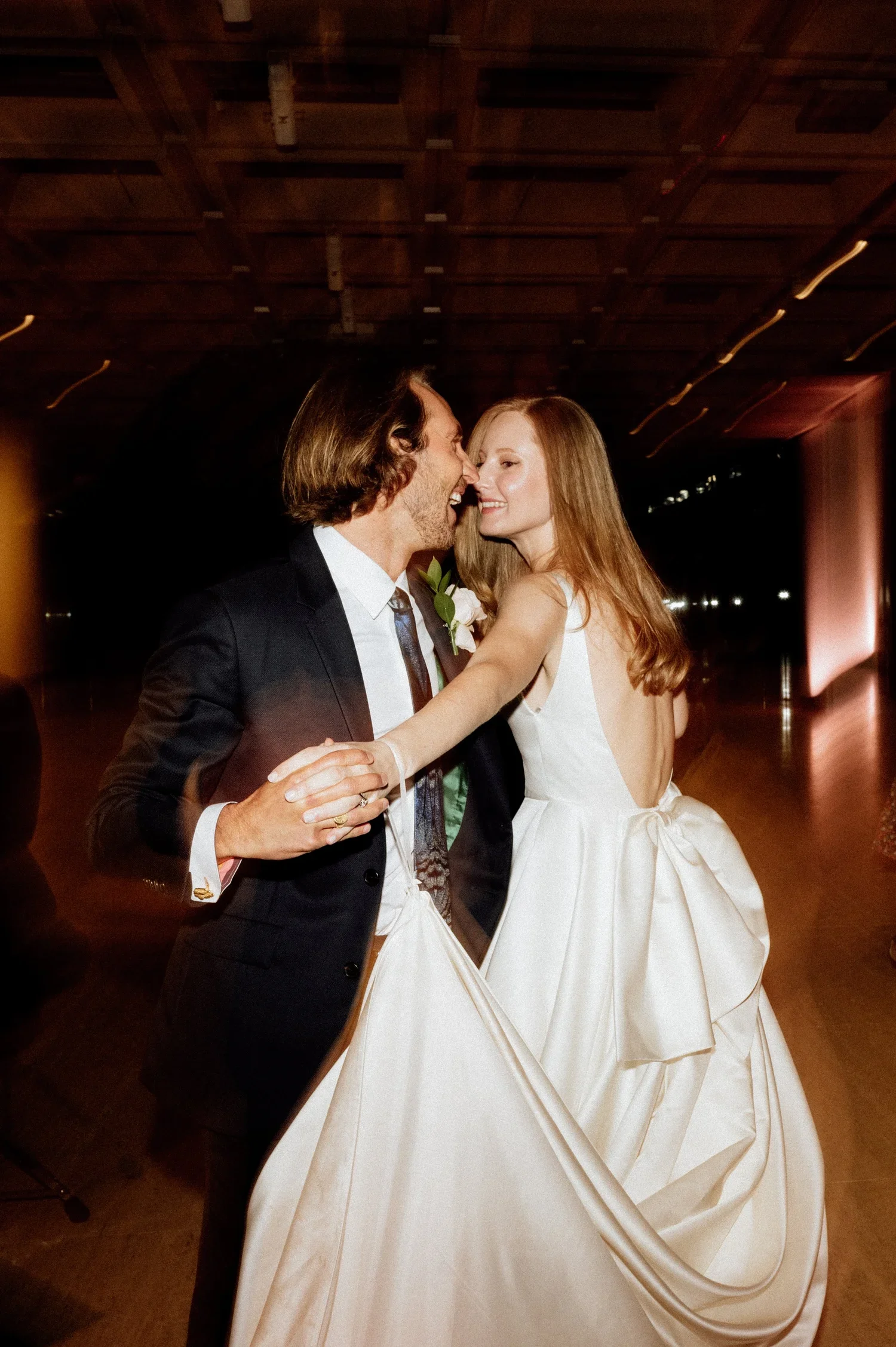 A couple dancing closely at a wedding reception, with the man wearing a black suit and tie, and the woman in a white wedding dress with a bow detail. They are smiling and looking at each other in a dimly lit venue with a wooden ceiling.