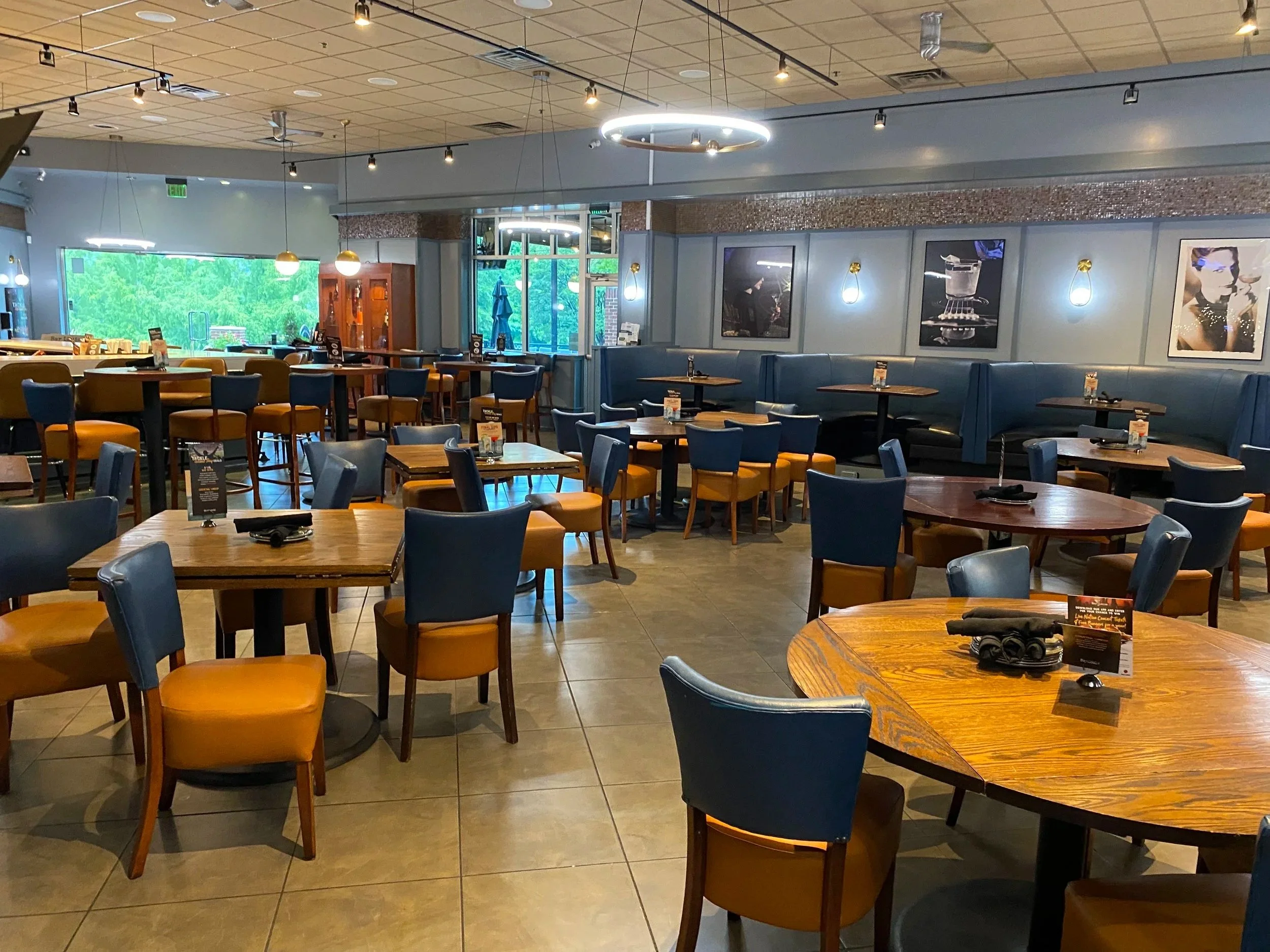 Interior of a modern restaurant with wooden tables, blue and mustard-colored chairs, decorated walls with framed art, and large windows letting in natural light.