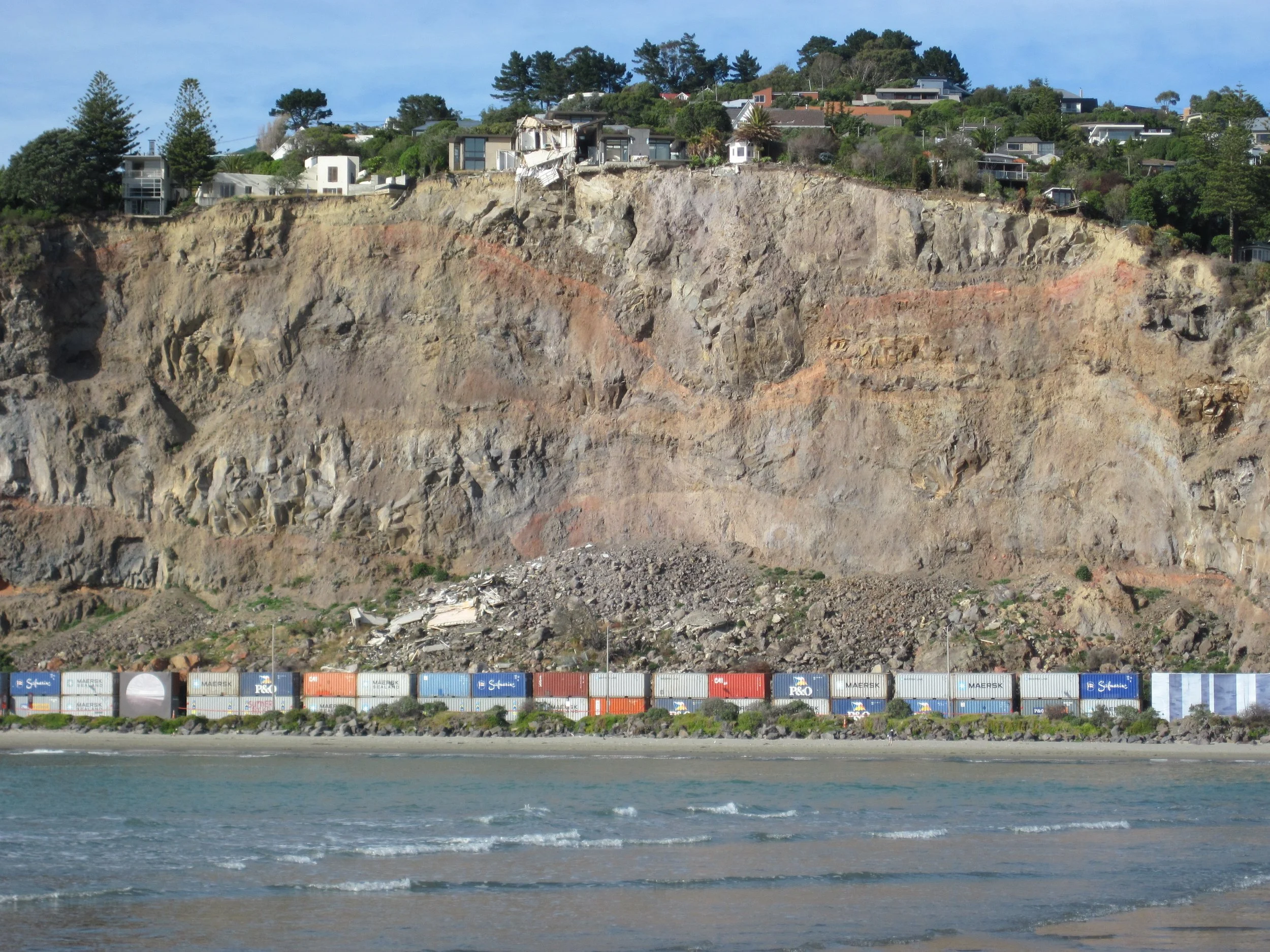 Image of a sheer cliff-face that has experienced a major landslide with a couple of houses dangling over it. and shipping containers creating a barrier at the bottom near the sea.