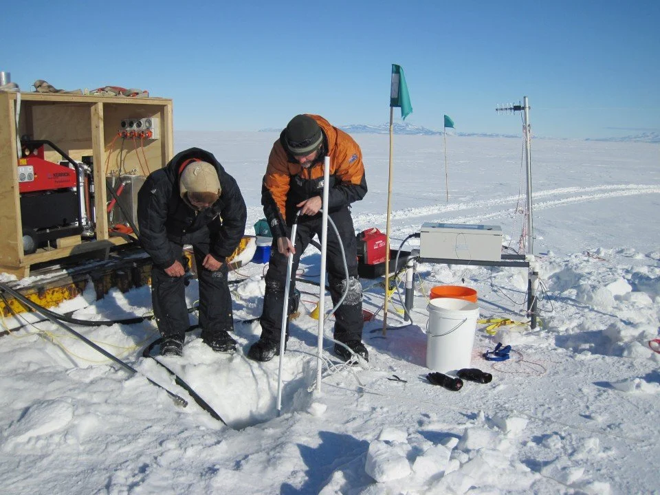 Scientists in cold weather gear conducting research in snow-covered terrain with equipment and flags.
