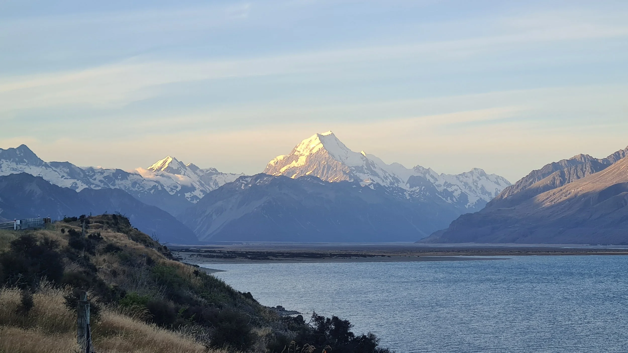 Mountain range with snow-capped peaks, a body of water in the foreground, and a partly cloudy sky.