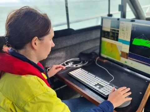 A woman working at a computer station with multiple screens displaying data, charts, and a 3D model of a green vehicle design.