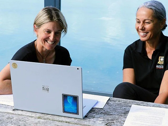 Two women with grey hair smiling and looking at a laptop on a wooden table, with a body of water in the background.