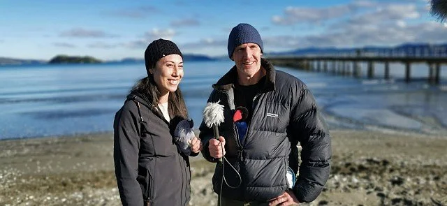 Two people standing on a beach near the water, dressed warmly in jackets and knit hats, with a pier in the background.