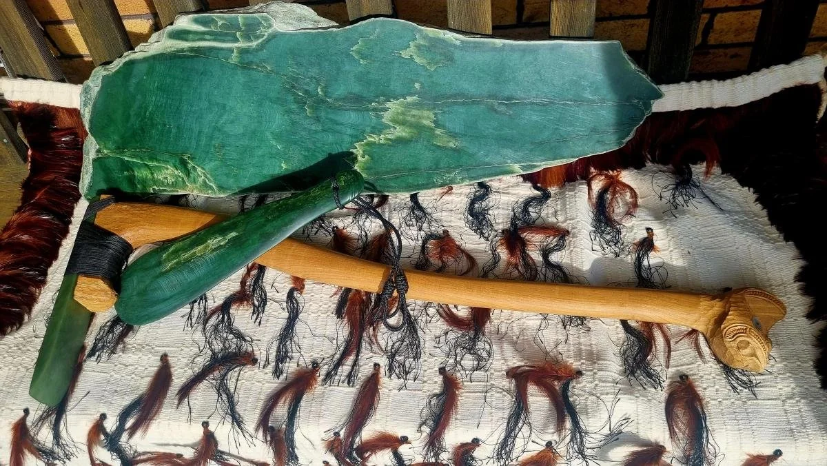 A split raw pounamu rock displayed with a mere pounamu, and a pounamu adze on top of a korowai.