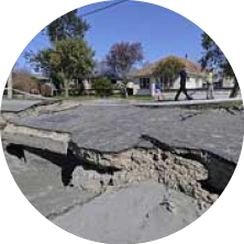 A cracked and damaged asphalt street with visible underlying gravel, trees, and houses in the background.
