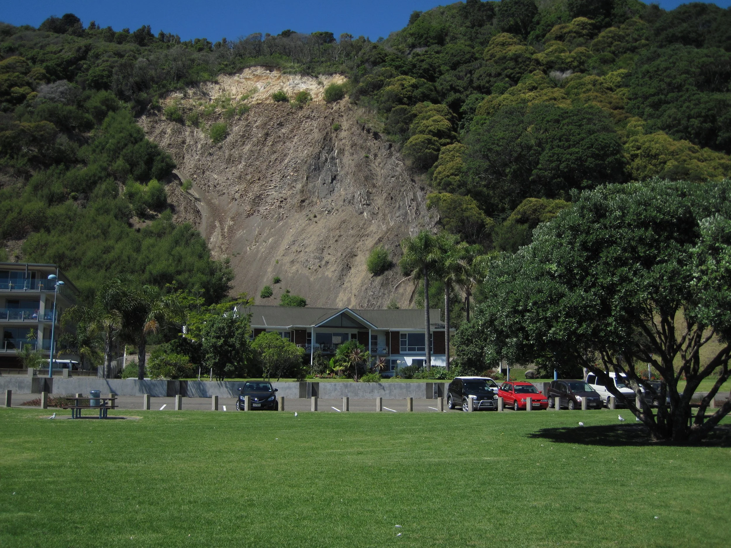 A hill showing a landslide with a car park and buildings in the foreground.