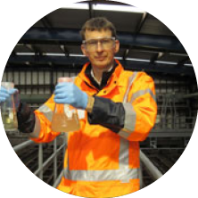 Young man wearing safety glasses, orange safety jacket, and blue gloves, holding a beaker with an orange liquid in a laboratory or industrial setting.