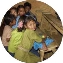Group of children gathered around a piano, with one girl sitting at the keyboard, in a classroom or indoor setting.