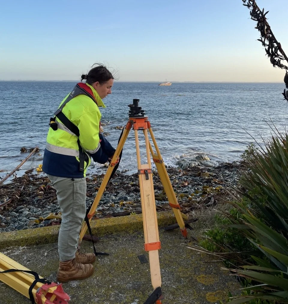 A woman wearing a hi-vis jacket standing at a beach with a theodolite tripod..